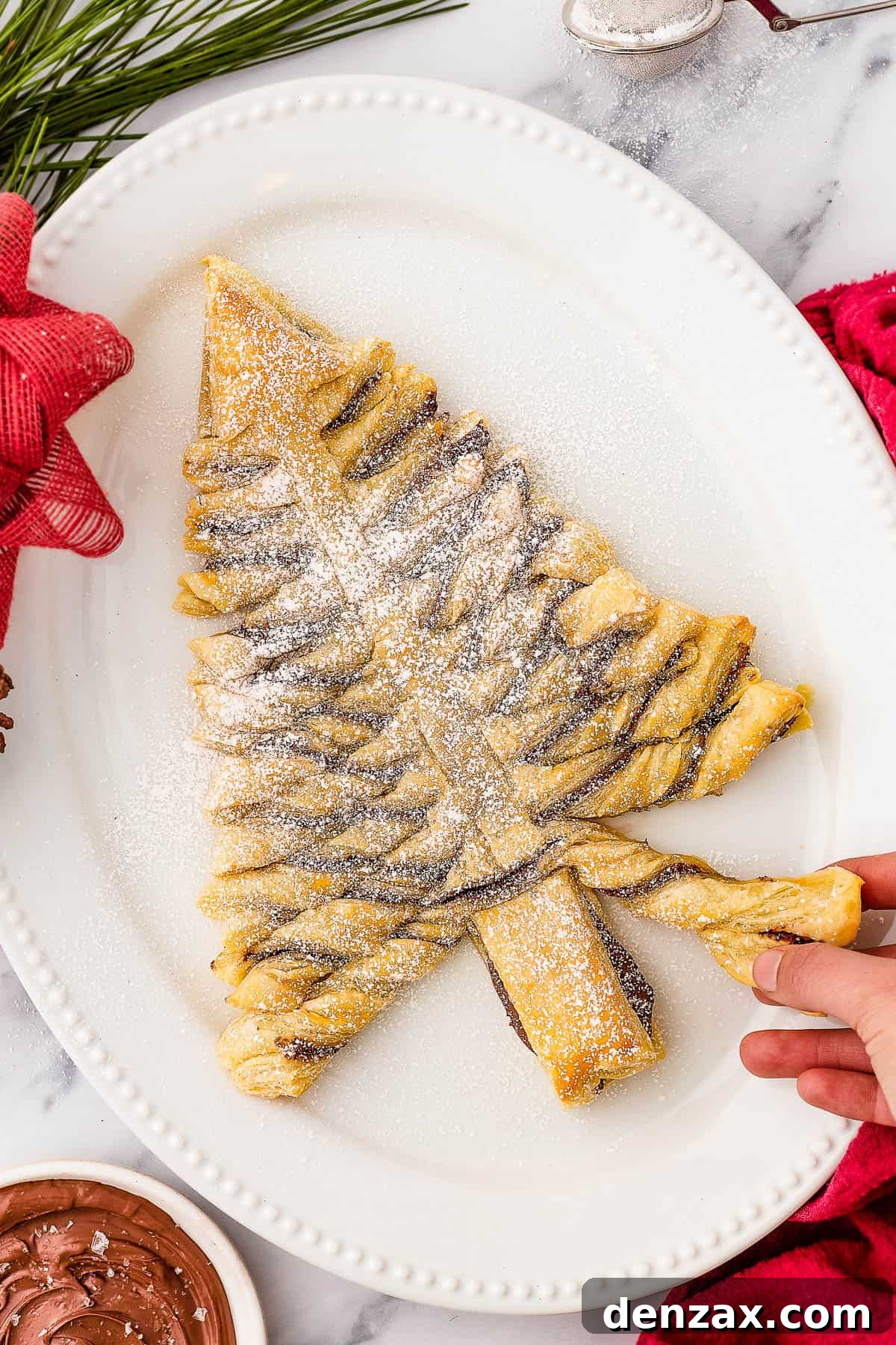 A baked puff pastry Christmas tree, dusted with powdered sugar, on an oval platter. Christmas greenery, a red bow, and a small bowl of chocolate dipping sauce surround the platter.