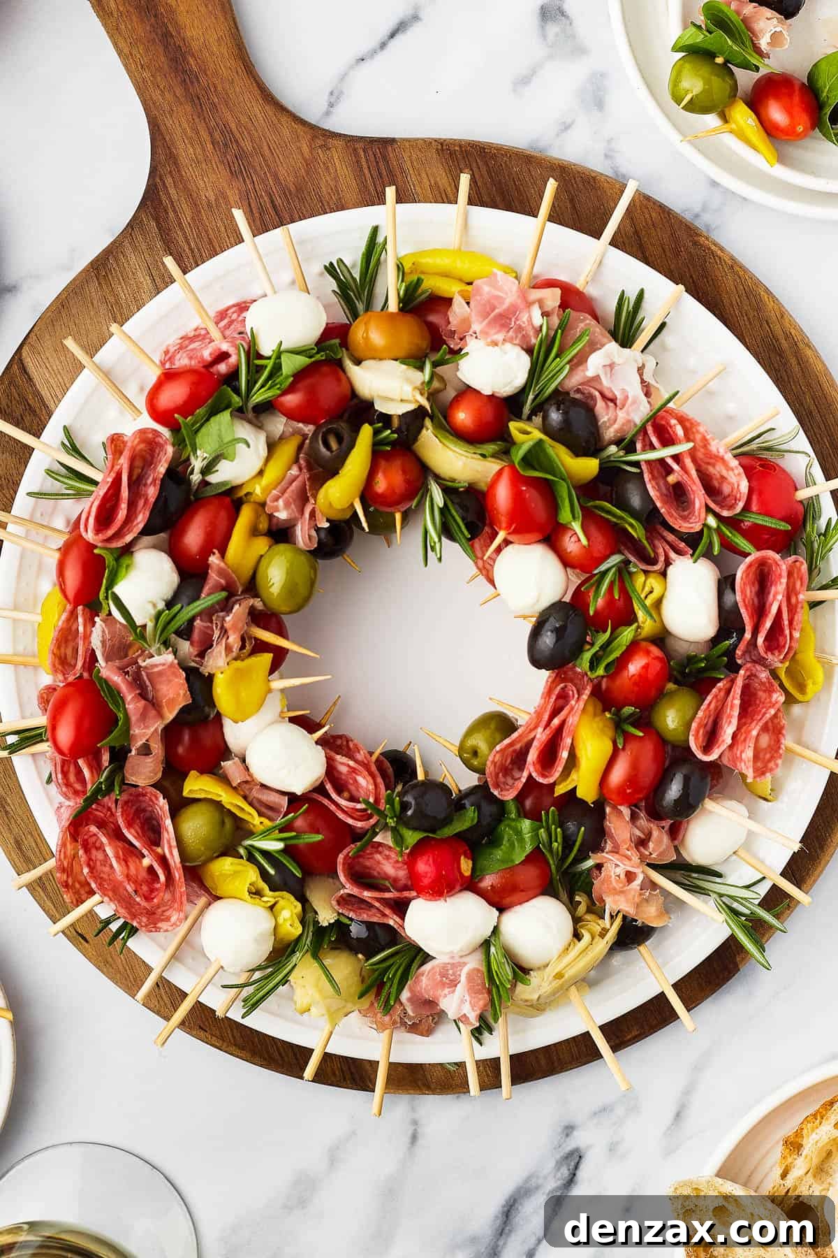 A table with a cutting board holding a wreath of skewers, a small pair of scissors, and fresh rosemary sprigs.