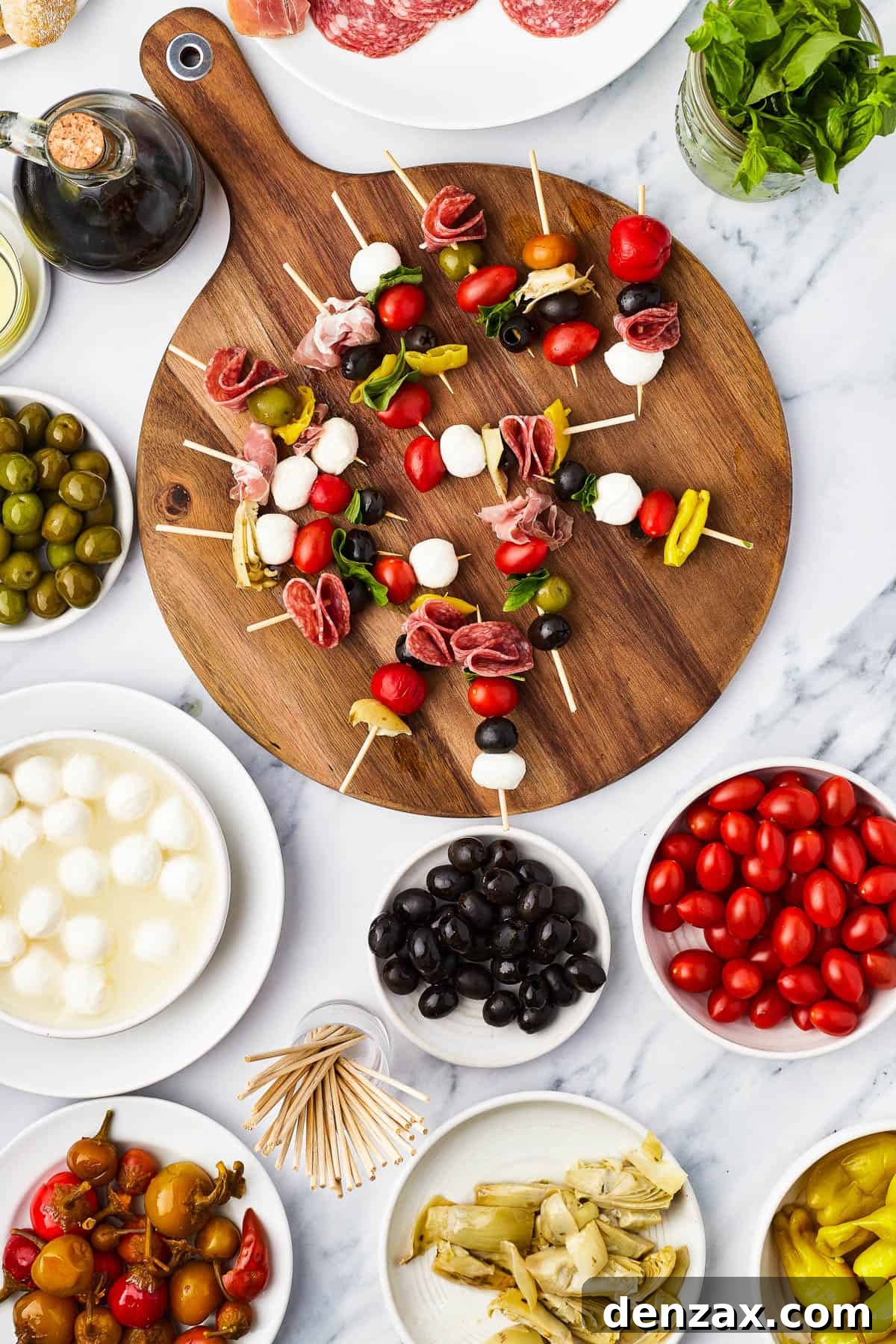 Skewers of olives, mozzarella, etc. on a round cutting board, surrounded by bowls of antipasto ingredients.