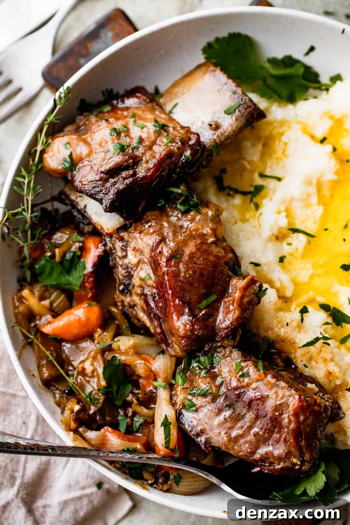 close up shot of a dinner plate with three short ribs arranged around mashed potatoes, with veggies and onions set on the other side of the short ribs.