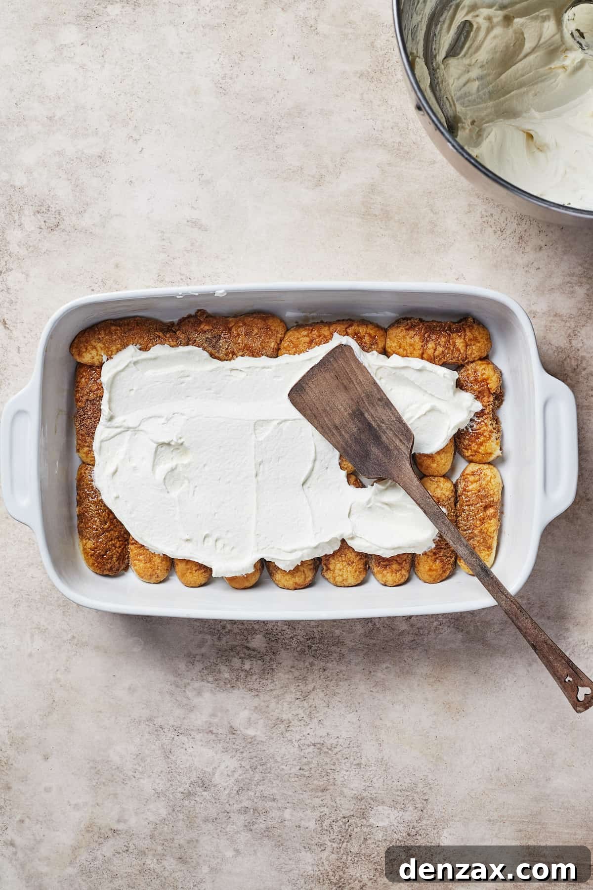 Creamy topping partially spread across a layer of savoiardi in a baking dish.
