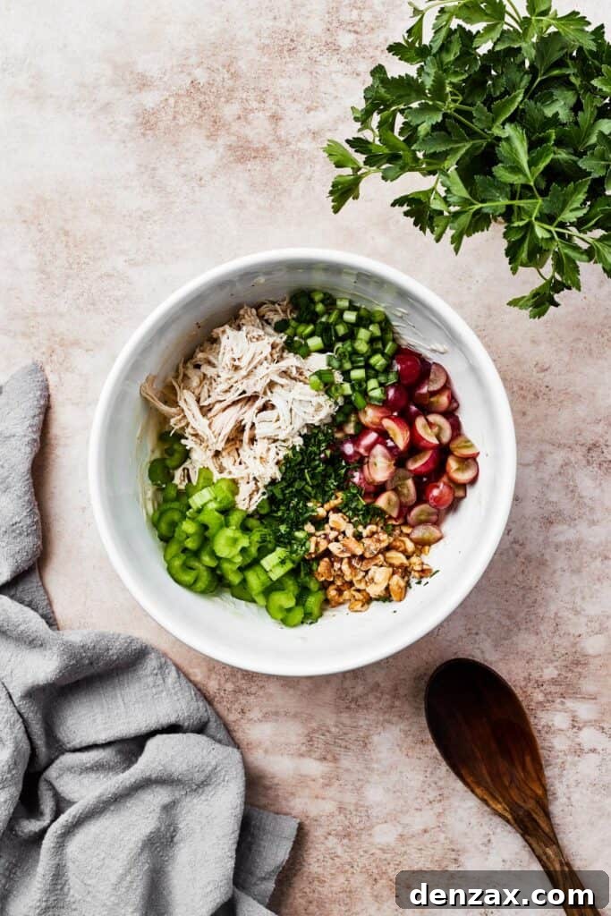 A white mixing bowl with shredded chicken, vegetables, grapes, and other ingredients.