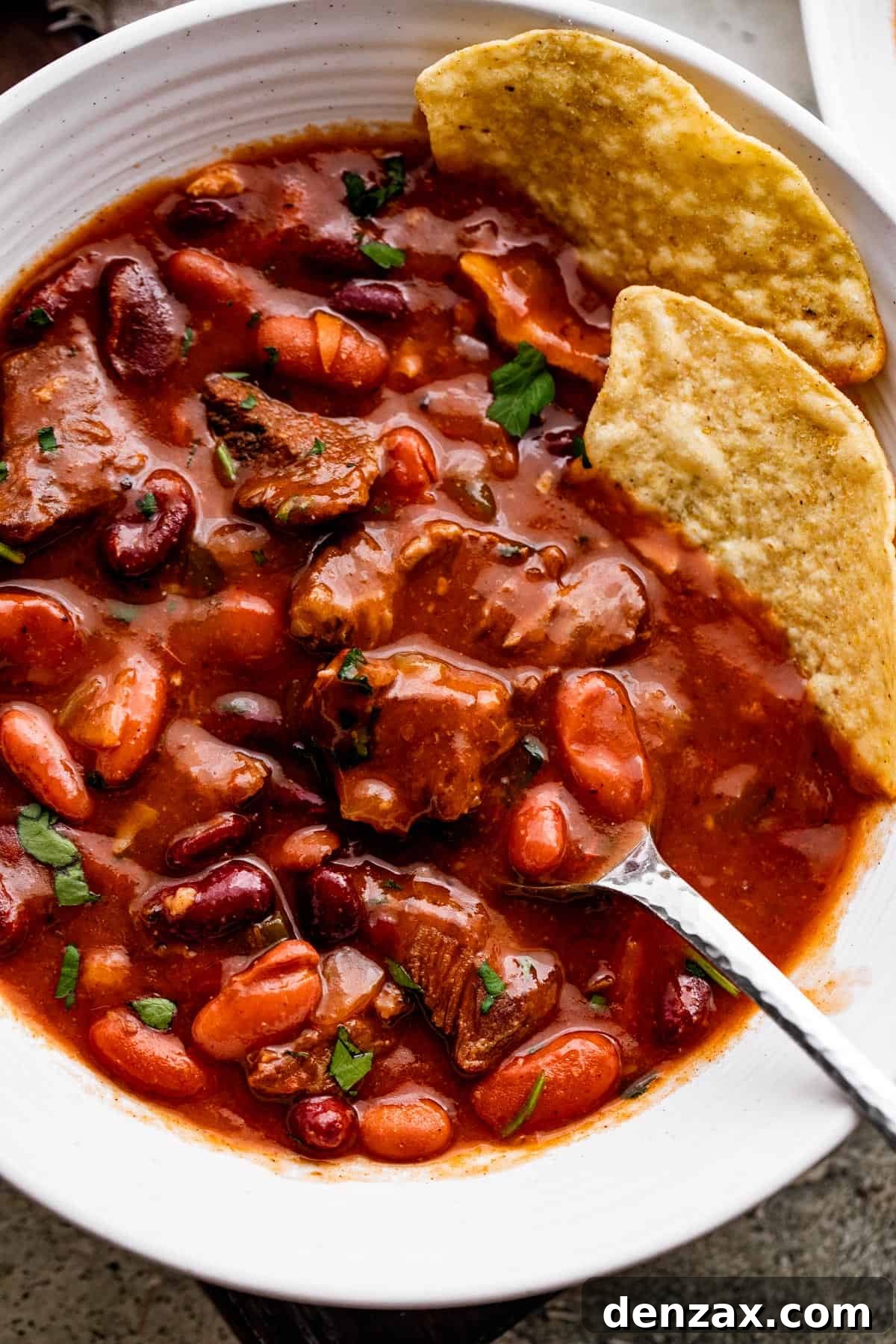 close up overhead shot of chili con carne in a white bowl, with a spoon and two tortilla chips set inside the chili