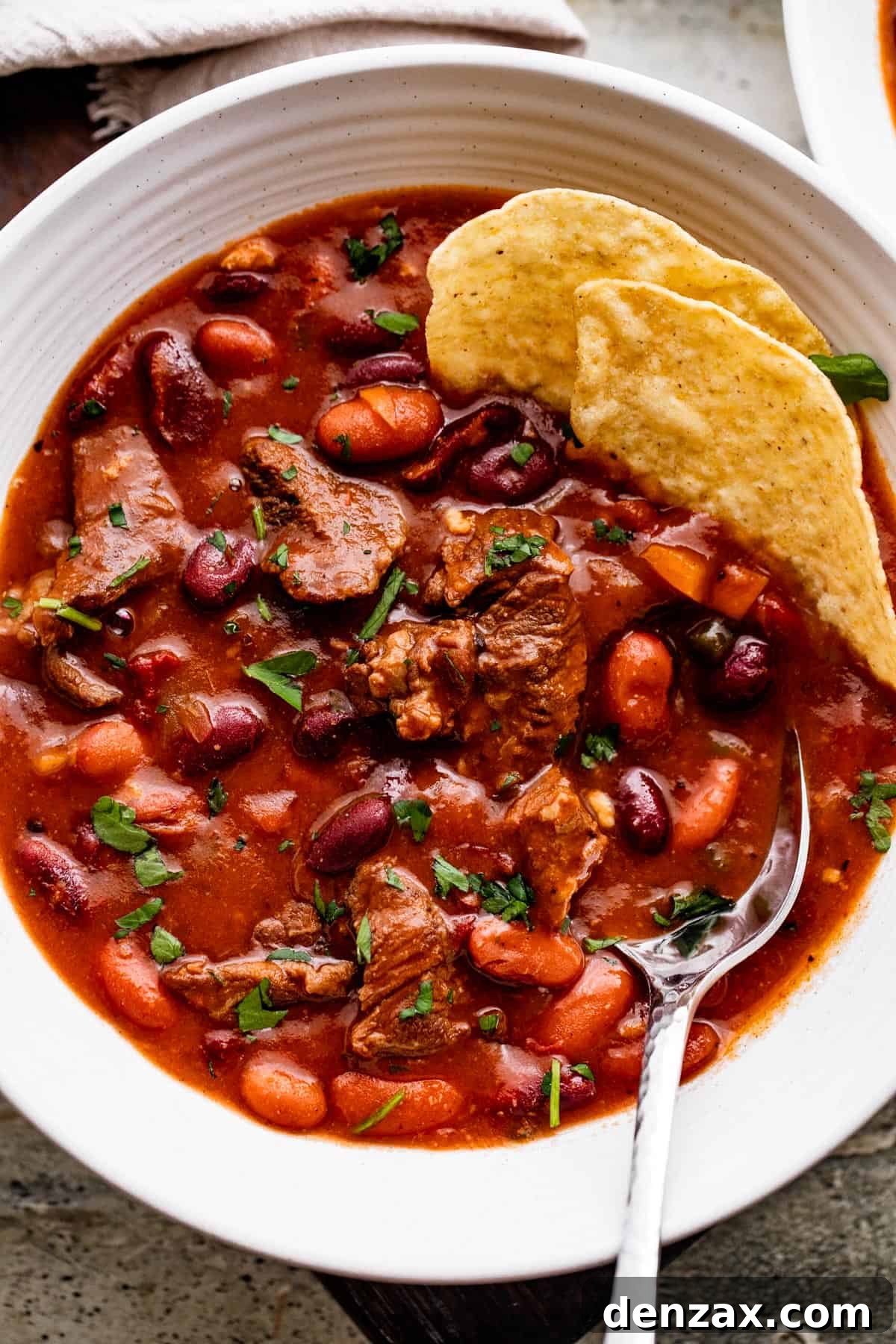 overhead shot of chili con carne in a white bowl, with a spoon and tortilla chips set inside the chili