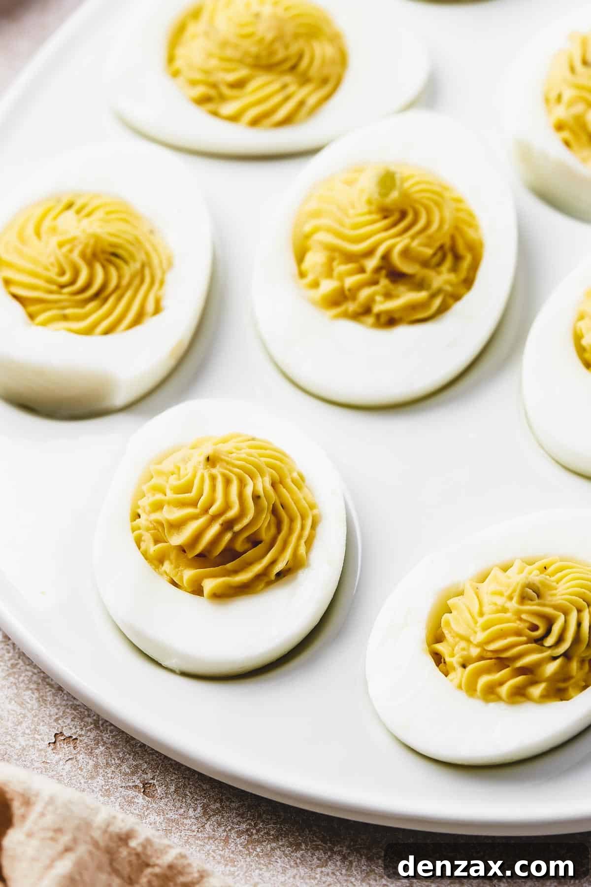 Classic Deviled Eggs Recipe: Creamy, Tangy Party Appetizer 2 A shallow white platter, shaped like an egg, with deviled eggs arranged on it. Bowls of garnishes are arranged around the platter.
