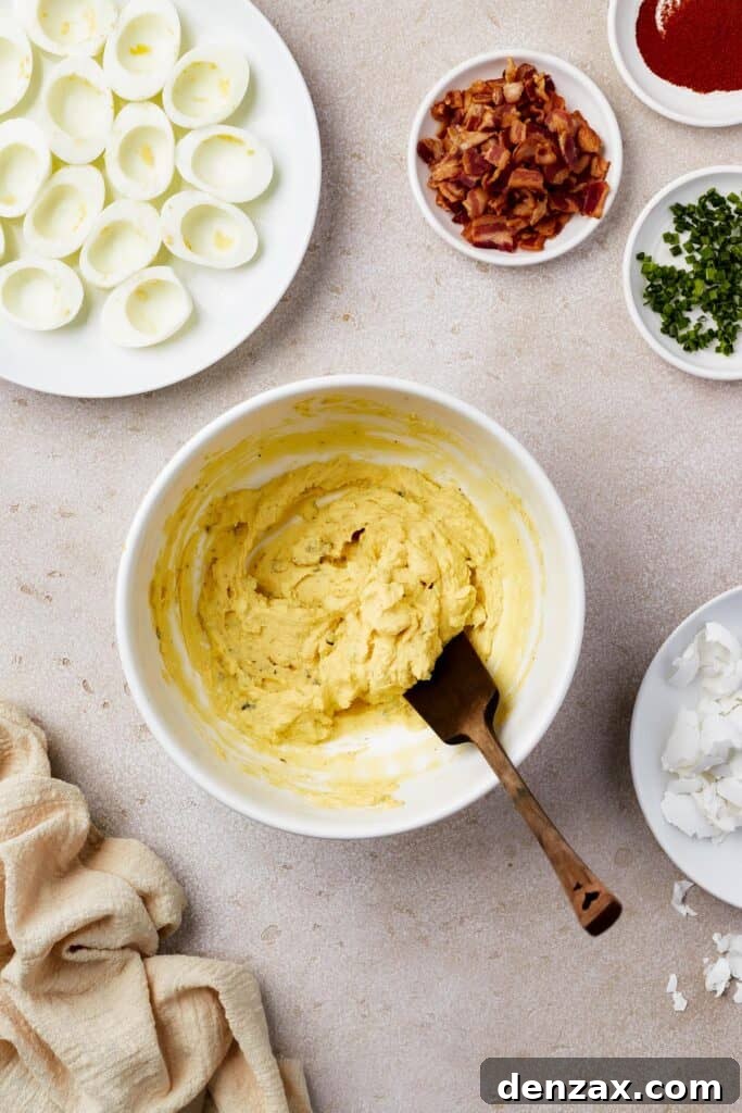 Classic Deviled Eggs Recipe: Creamy, Tangy Party Appetizer 6 A bowl of creamy egg yolk filling with a wooden spoon. Cooked egg whites, bacon, and other ingredients are on the work surface also.