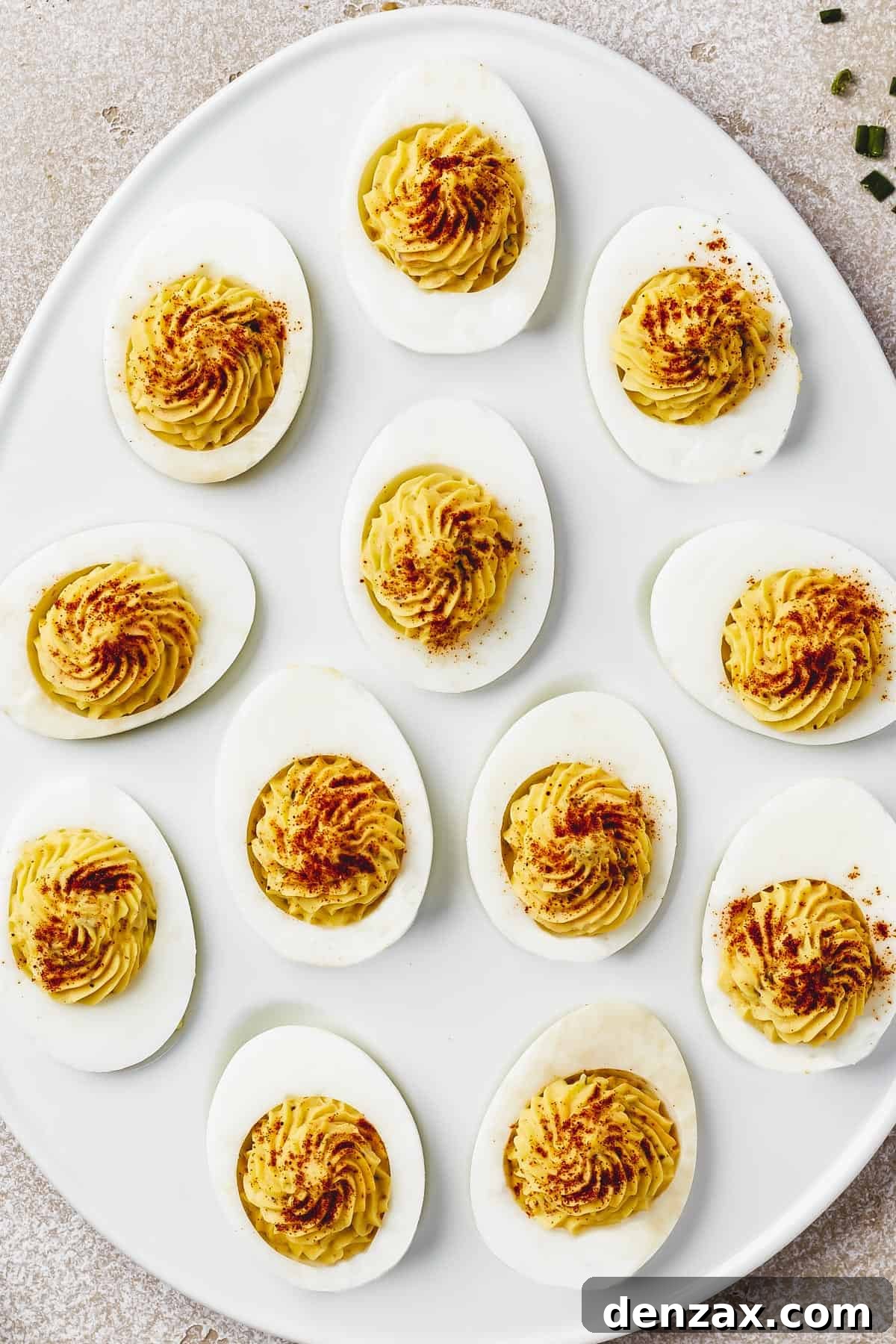Classic Deviled Eggs Recipe: Creamy, Tangy Party Appetizer 9 Overhead shot of an egg-shaped white dish with stuffed eggs arranged on it. Each egg has been garnished with paprika, bacon, and chives.