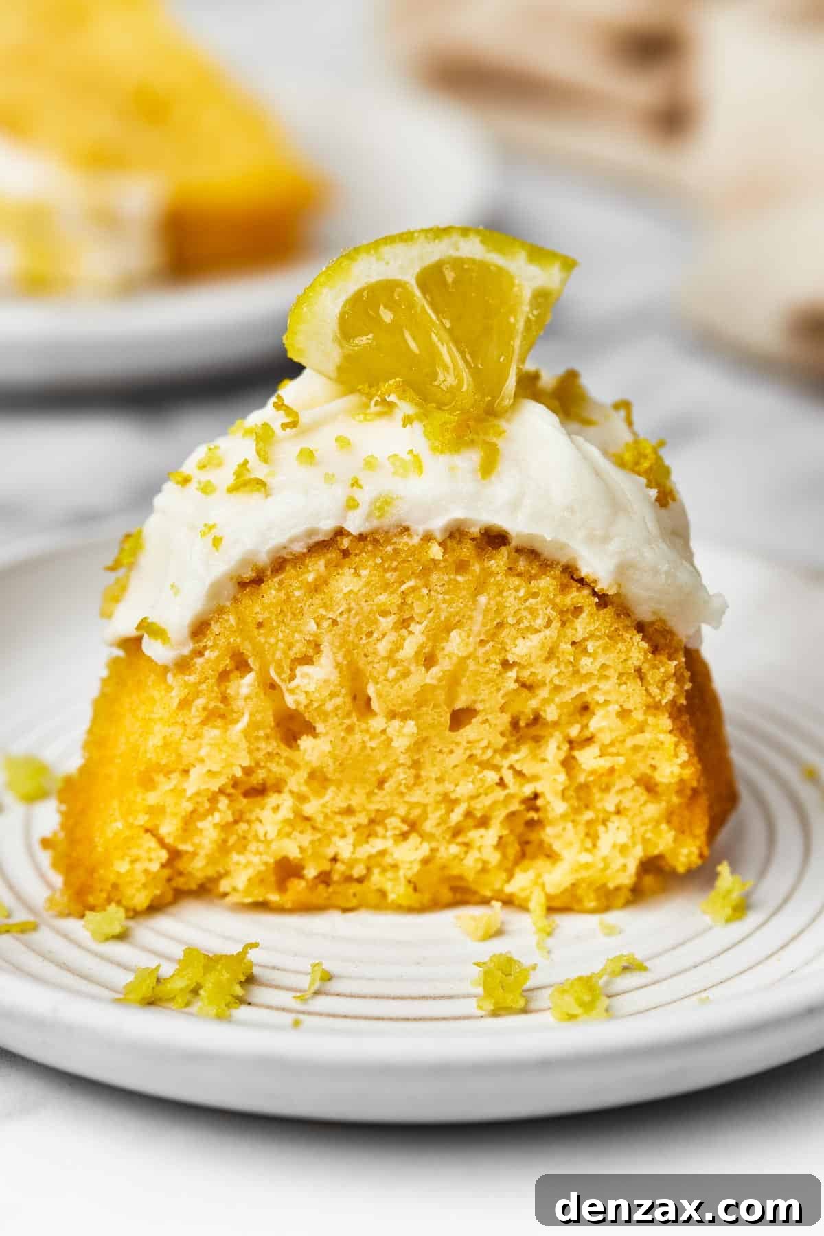 A slice of frosted cake, garnished with lemon zest and a small lemon wedge.
