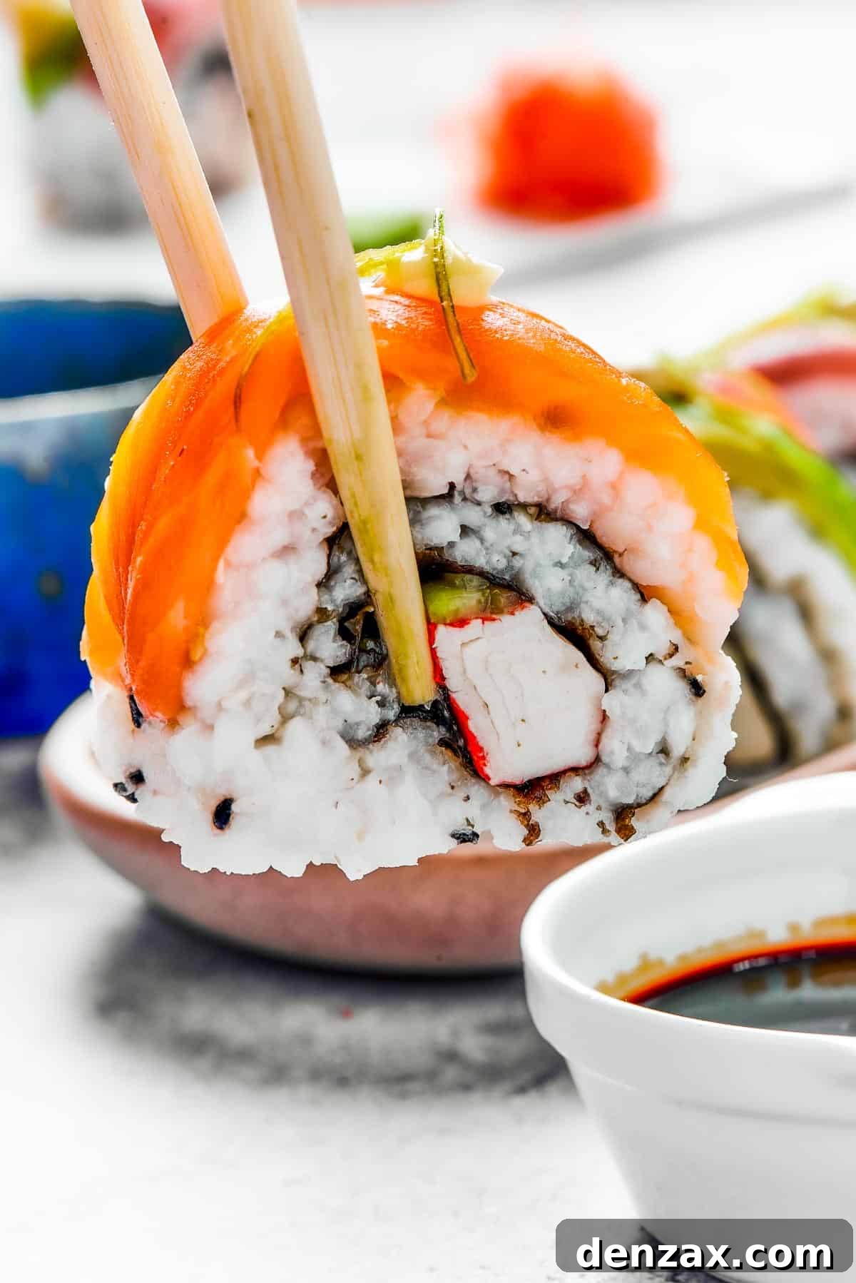 A bite of sushi held in a pair of chopsticks. A small plate of sushi and a container of soy sauce is in the background.