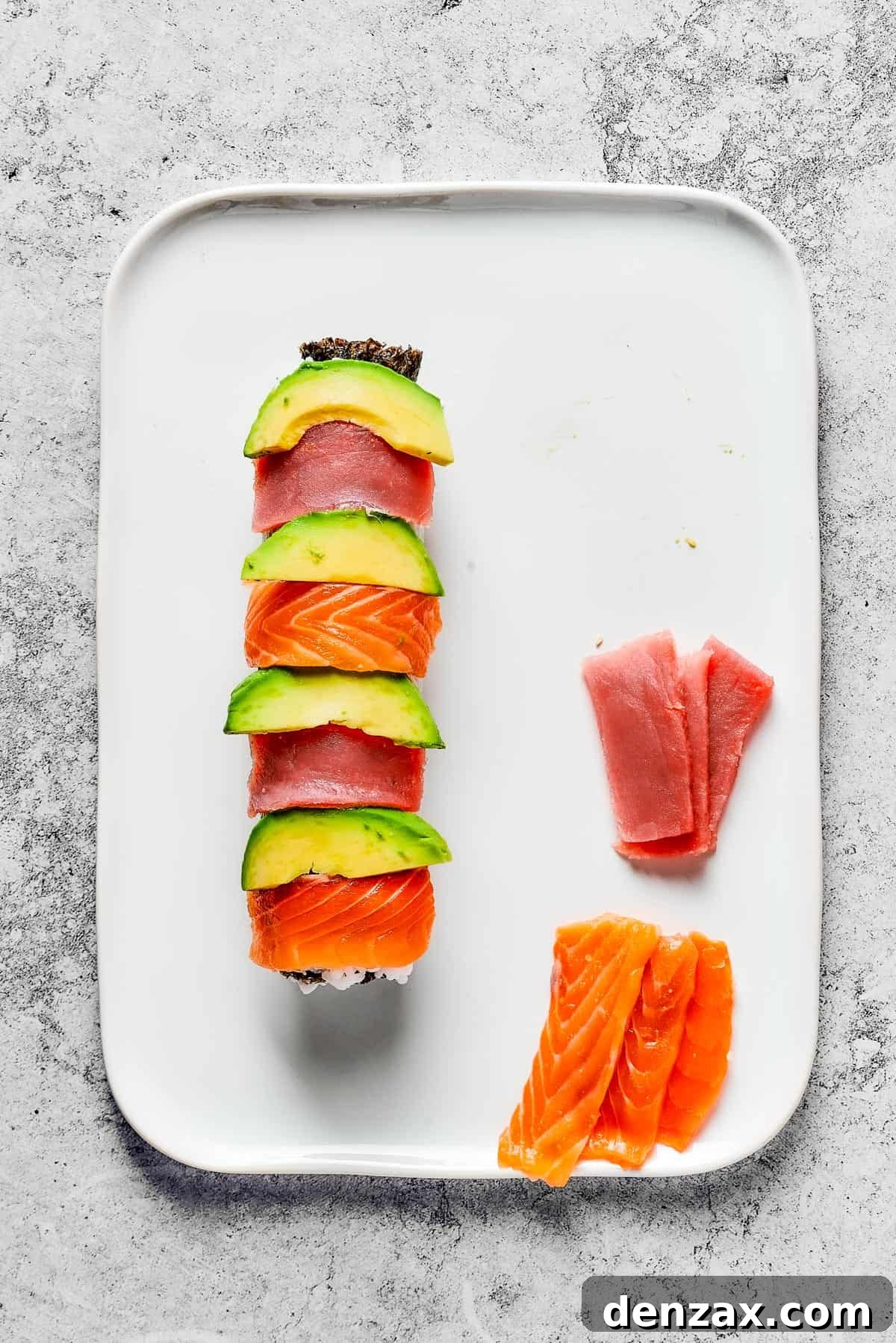 Overhead shot of a roll of sushi, topped with alternating slices of avocado, tuna, and salmon. Extra slices of tuna and salmon are on the platter.