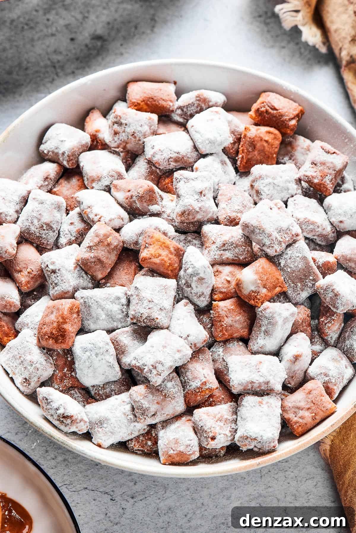 Close-up shot of puppy chow in a white bowl, to show the texture. Some pieces are fully coated in powdered sugar, while others are not.