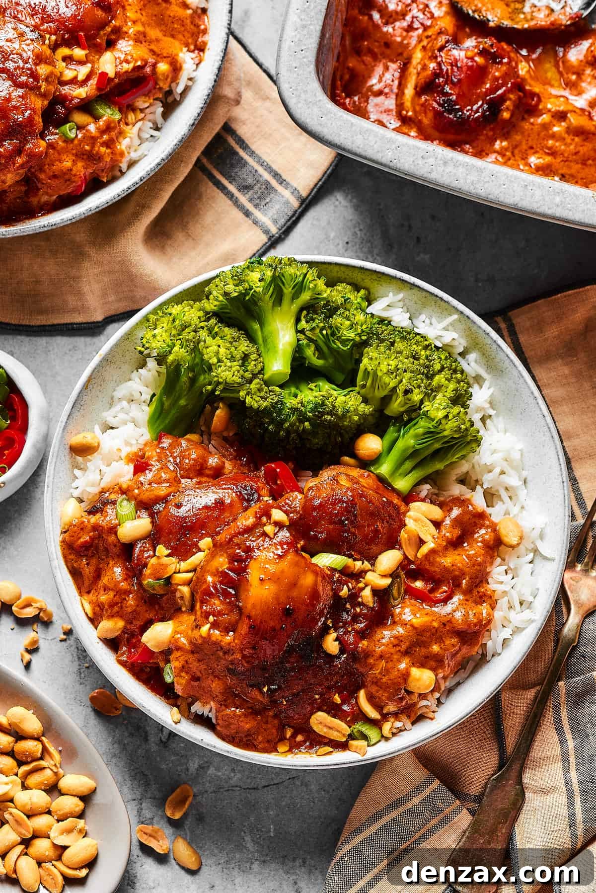 Peanut chicken served with broccoli over rice. A baking dish of the chicken is nearby on the table, along with garnishes and more plates.