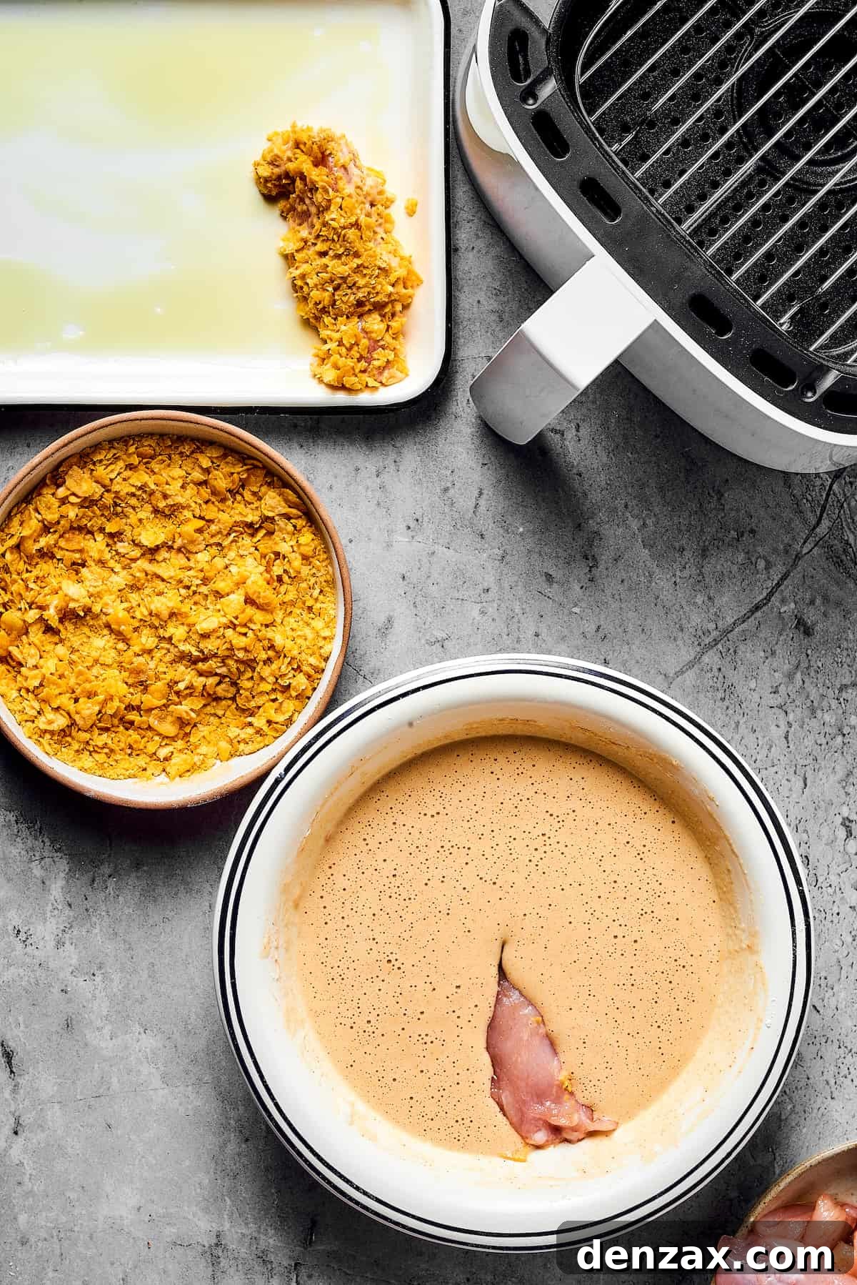 A piece of chicken dipped in batter, next to a bowl of cornflakes. A breaded chicken strip is on an oiled baking sheet nearby, along with an open air fryer.