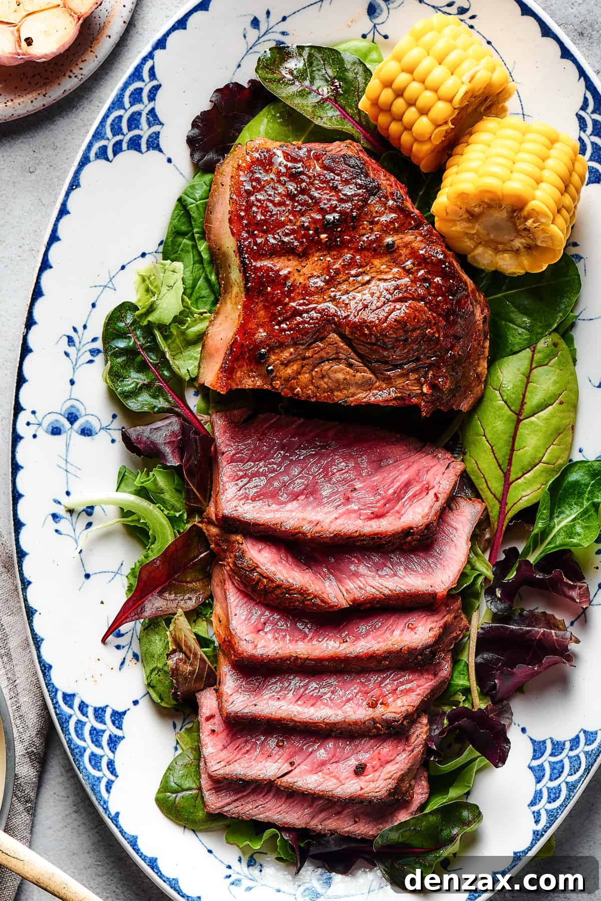 A steak on a platter of greens, garnished with corn on the cob. About half the steak has been sliced and fanned out to show the rare interior.