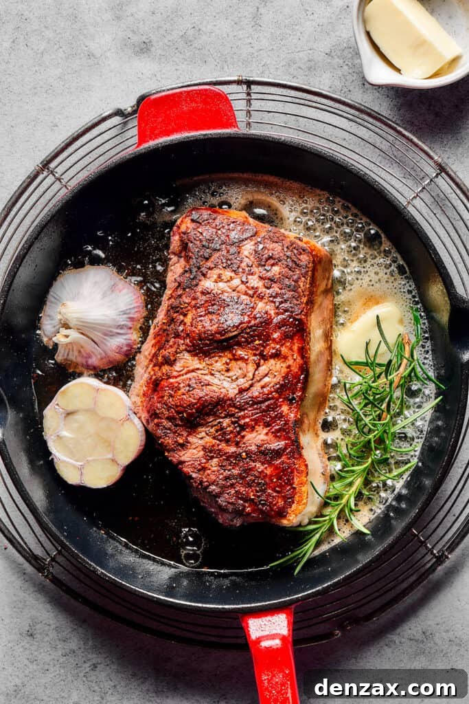 Sirloin steak in a cast-iron pan with melting butter, a sprig of rosemary, and a cut head of garlic.