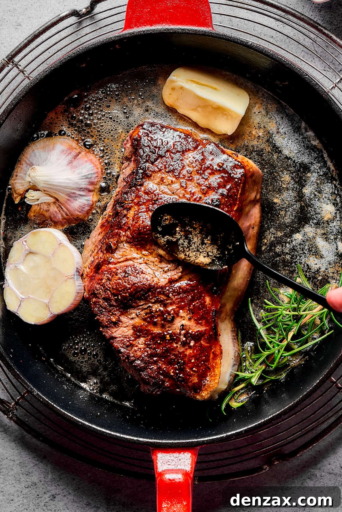 A person's hand basting pan juices over a steak, as the steak cooks in a skillet.
