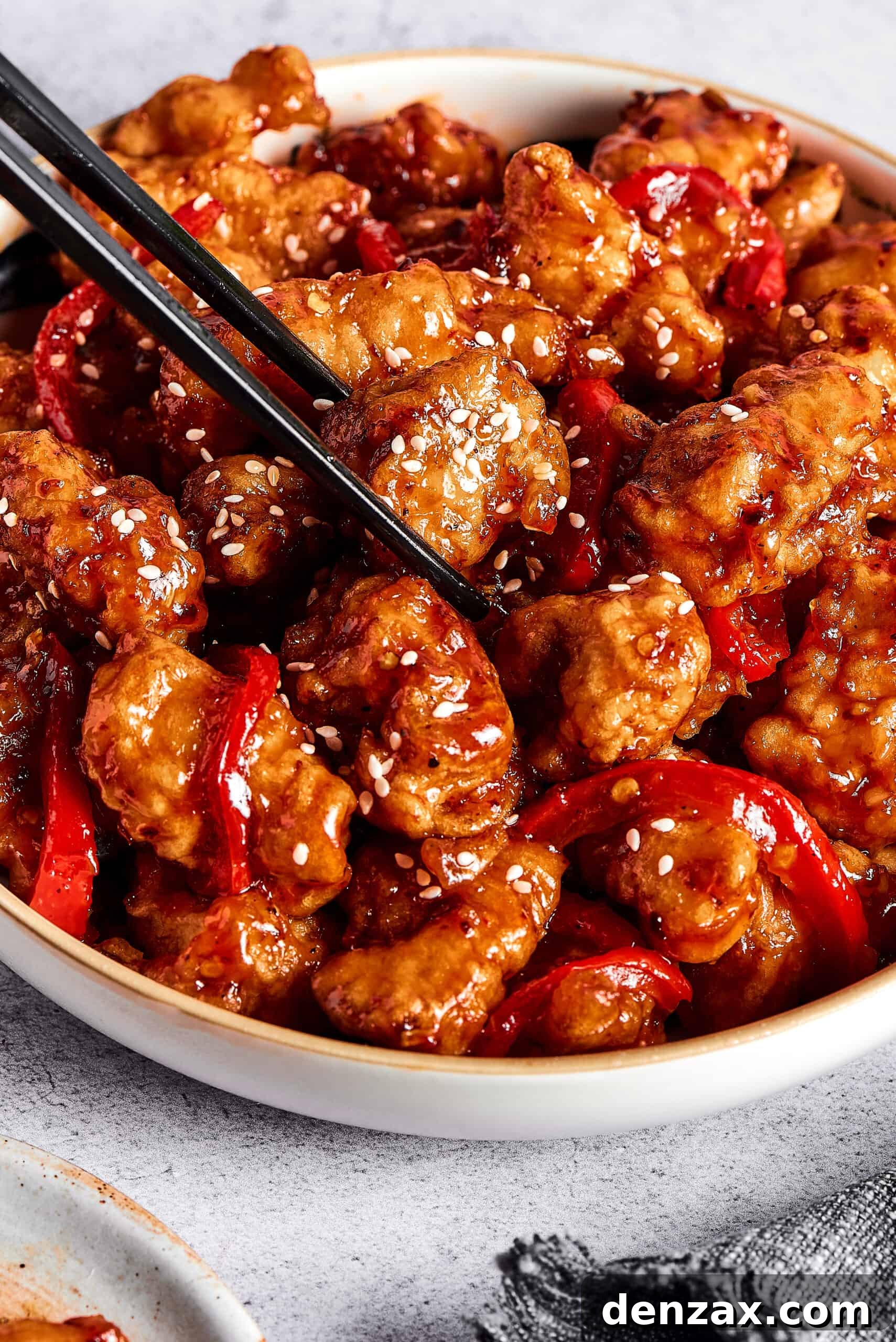 A pair of chopsticks picking up a piece of sticky chicken from a dish.