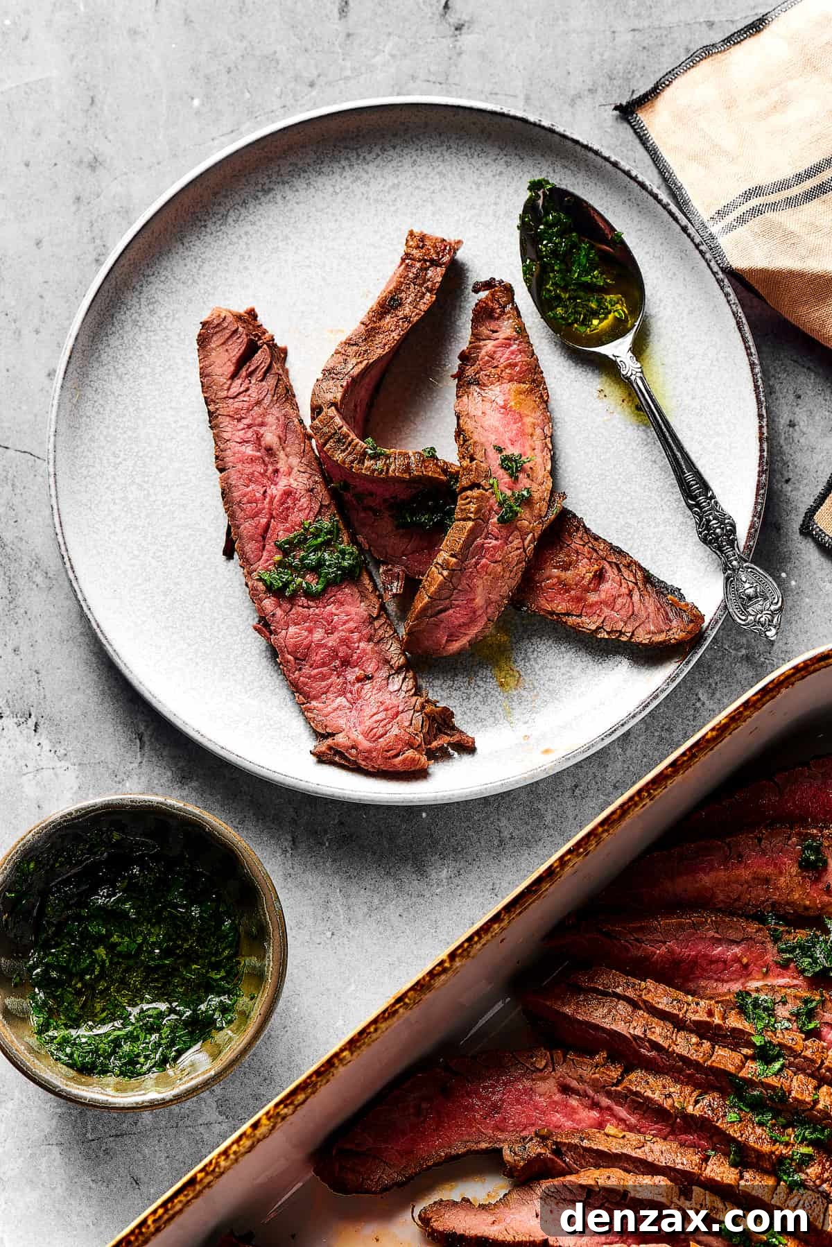 A small plate with three strips of medium-rare grilled carne asada on it. Nearby is a large platter of the steak slices, a cup of sauce, and a spoon.