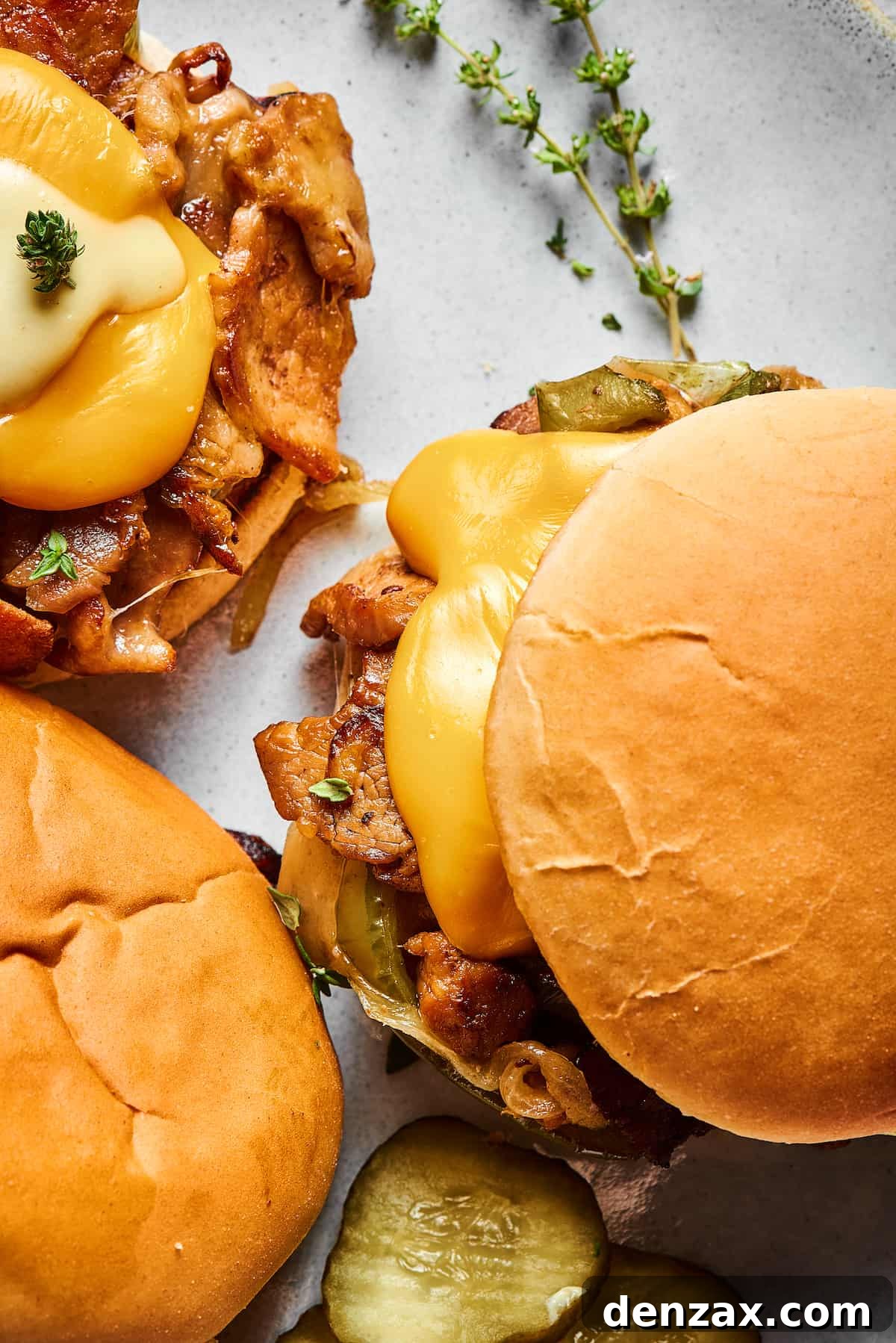Close-up shot of three philly cheesesteak burgers on a platter with dill pickle chips and a sprig of thyme.