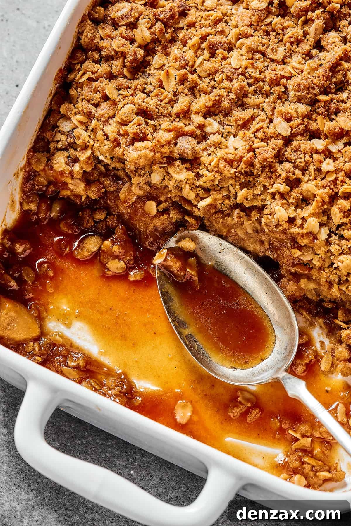 Close-up shot of the corner of a white casserole dish with an apple dessert, showing the apples, topping, and syrup. A spoon is resting in the empty part of the casserole dish.