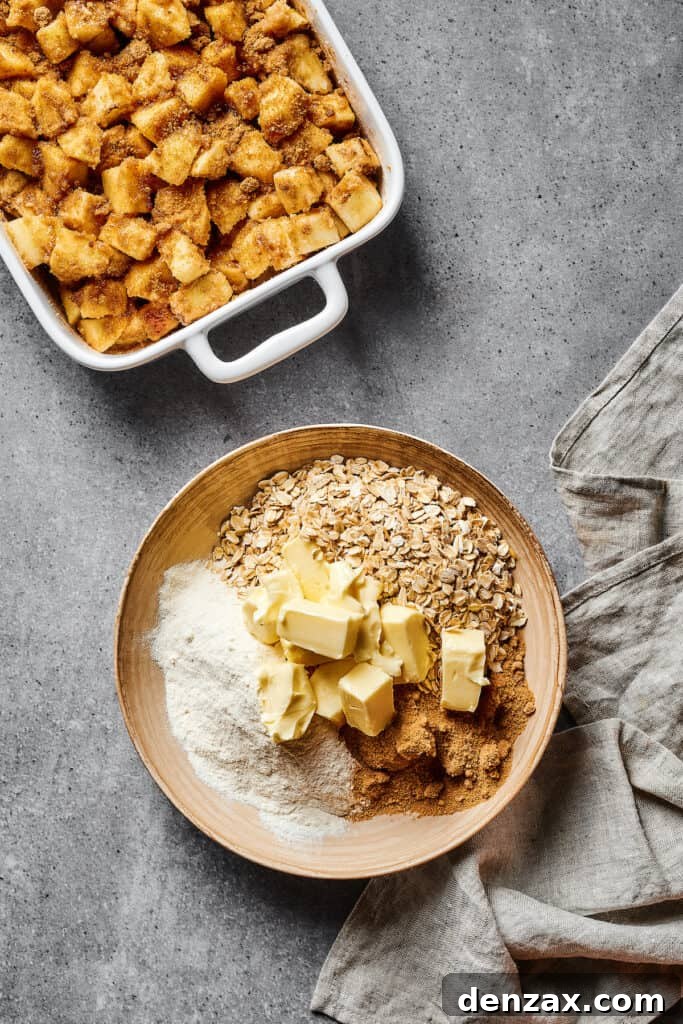 Spiced, floured apples in a casserole dish, next to a bowl of oats, cubed butter, and other baking ingredients.