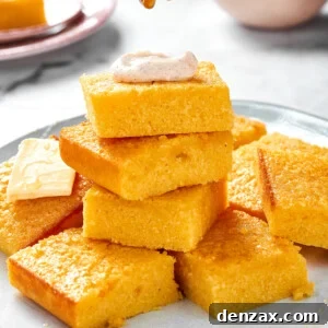 A platter of cornbread with some squares stacked on each other. The top piece of cornbread has a dollop of cinnamon cream on top and is being drizzled with honey.