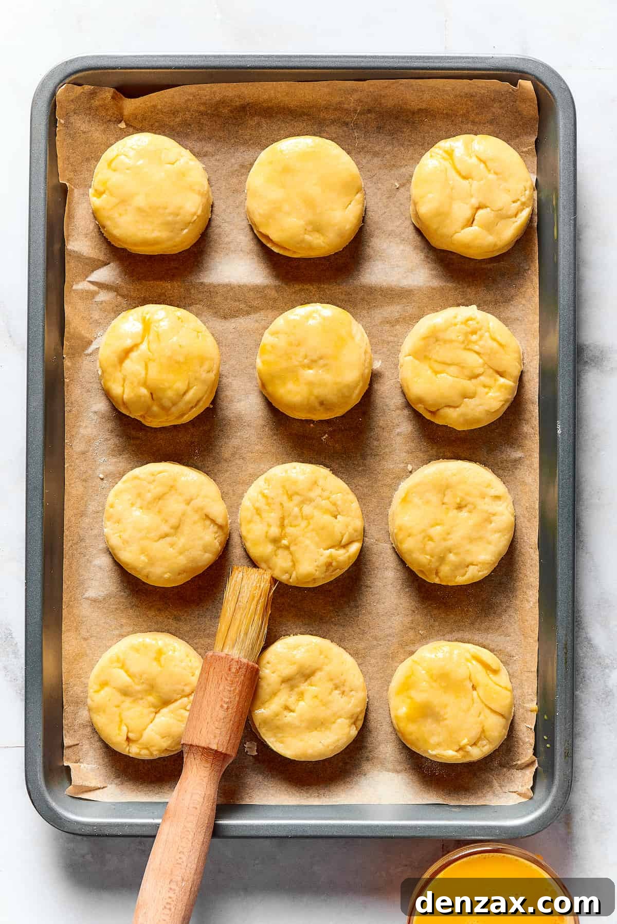 A dozen unbaked biscuits on a baking sheet, being brushed with beaten egg.