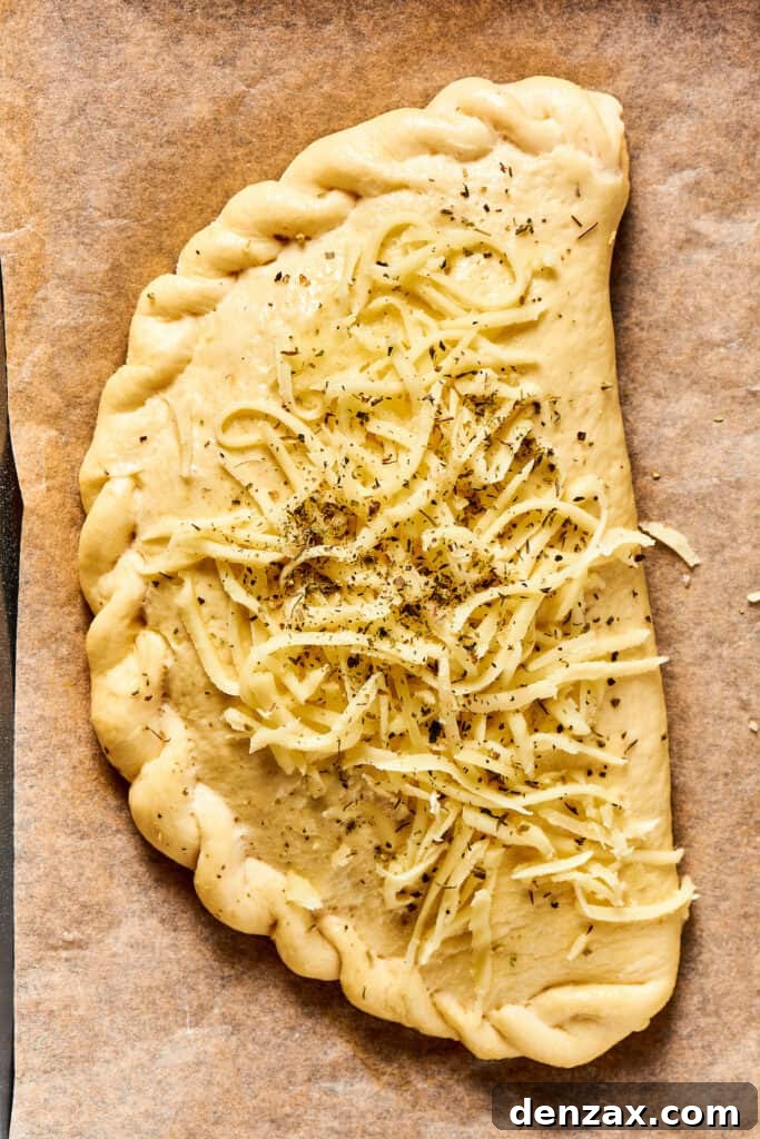 An unbaked calzone topped with shredded cheese and seasoning.