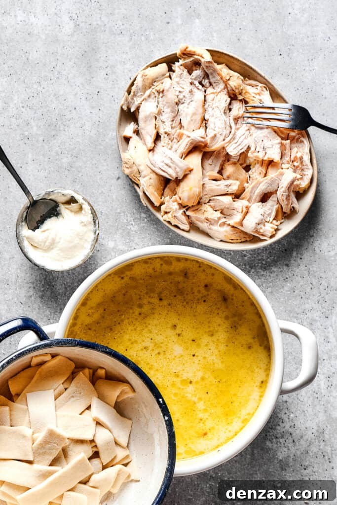 A pot of soup next to two dishes, one of shredded chicken and one of rolled dumplings. A measuring cup of thick cream is nearby on the work surface.