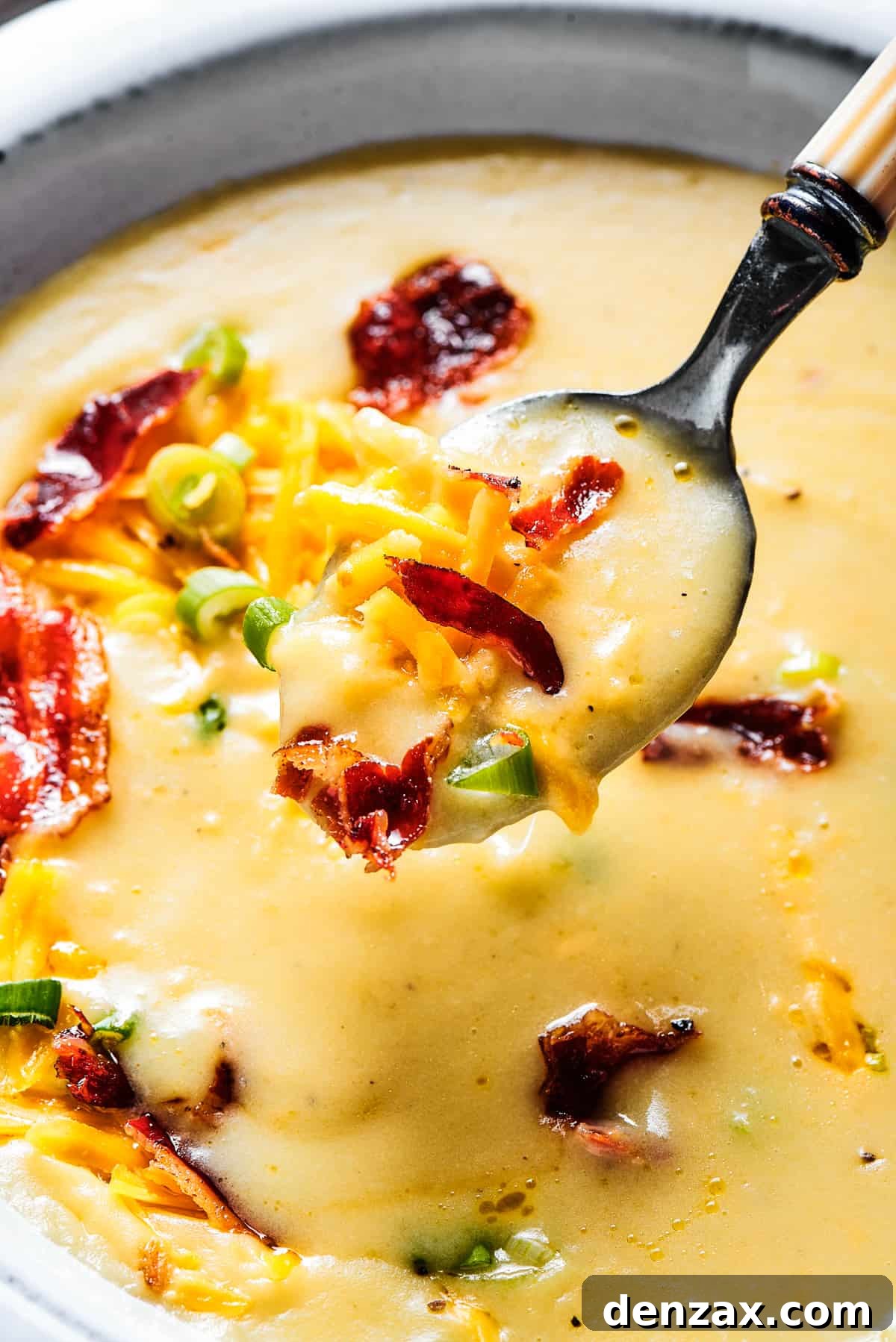 A white bowl of loaded potato soup, with a spoon lifted out of the bowl toward the camera.