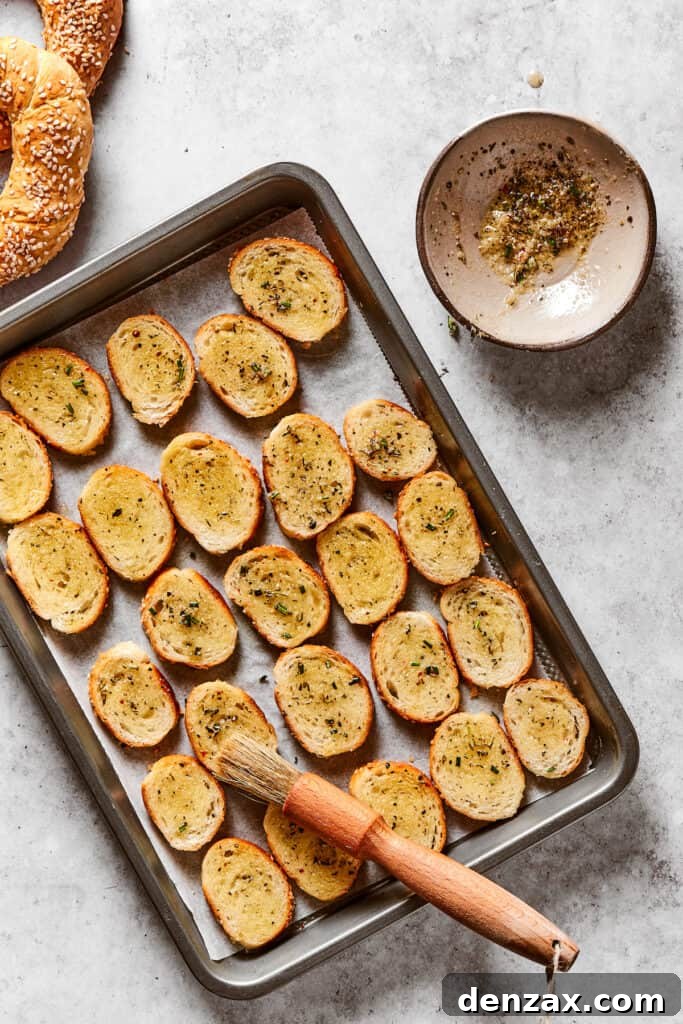 Bagel rounds lined up on a baking sheet, brushed with seasoned butter. A pastry brush is resting on the baking sheet, and dish of seasoned butter is next to it.