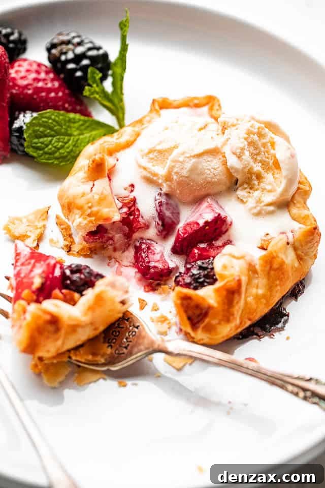 Mini berry galettes are the perfect summer dessert. Fresh berries are baked in a flaky pie crust that’s been dusted with sugar. So good! side view f berries stuffed in a mini galette and topped with ice cream