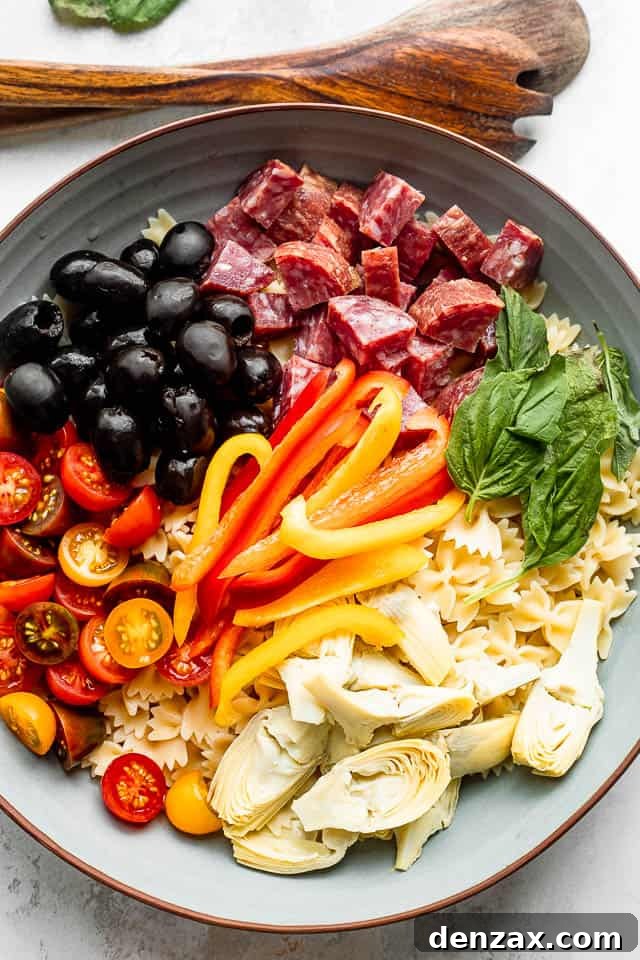 wooden spoons next to a salad bowl with arranged olives, salami, pasta, cheese, artichokes, tomatoes, basil leaves, and peppers