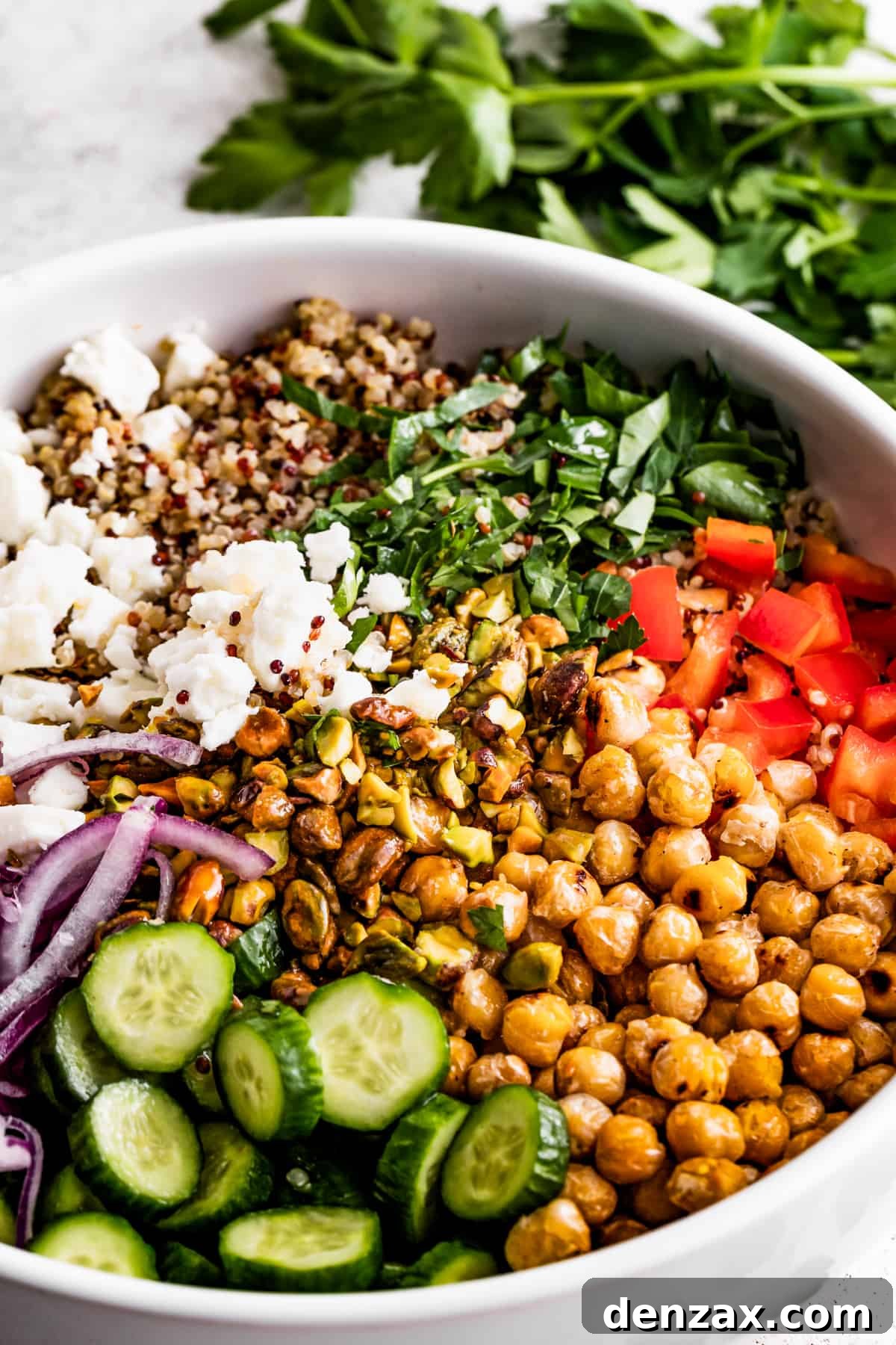 Drizzled with homemade dressing, this Jennifer Aniston quinoa salad with crunchy chickpeas is super light and refreshing. A large bowl with sliced cucumbers, sliced red onions, chickpeas, diced red bell peppers, herbs, tri-colored quinoa, and crumbles of feta cheese.