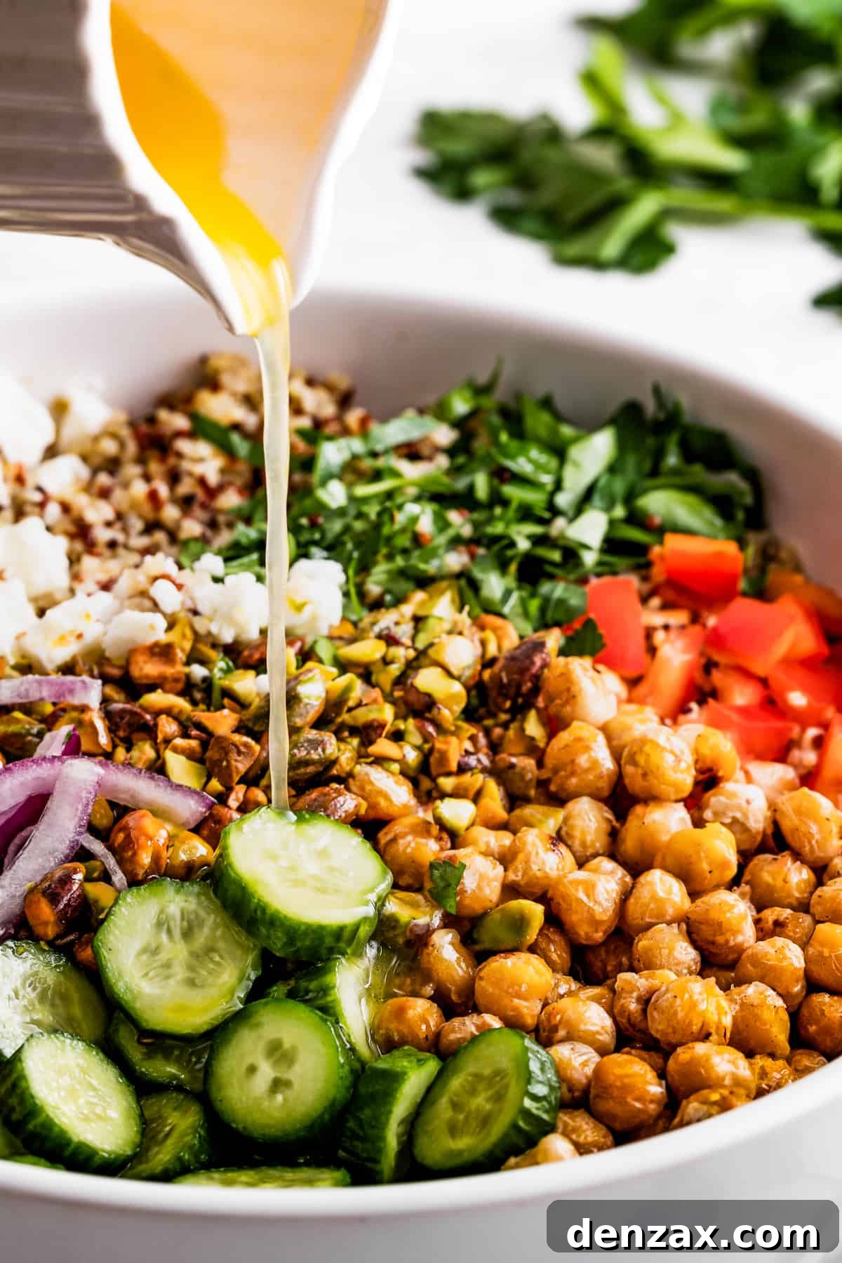 Drizzled with homemade dressing, this Jennifer Aniston quinoa salad with crunchy chickpeas is super light and refreshing. Pouring salad dressing over chickpeas, sliced cucumbers, red onions, peppers, herbs, and feta.