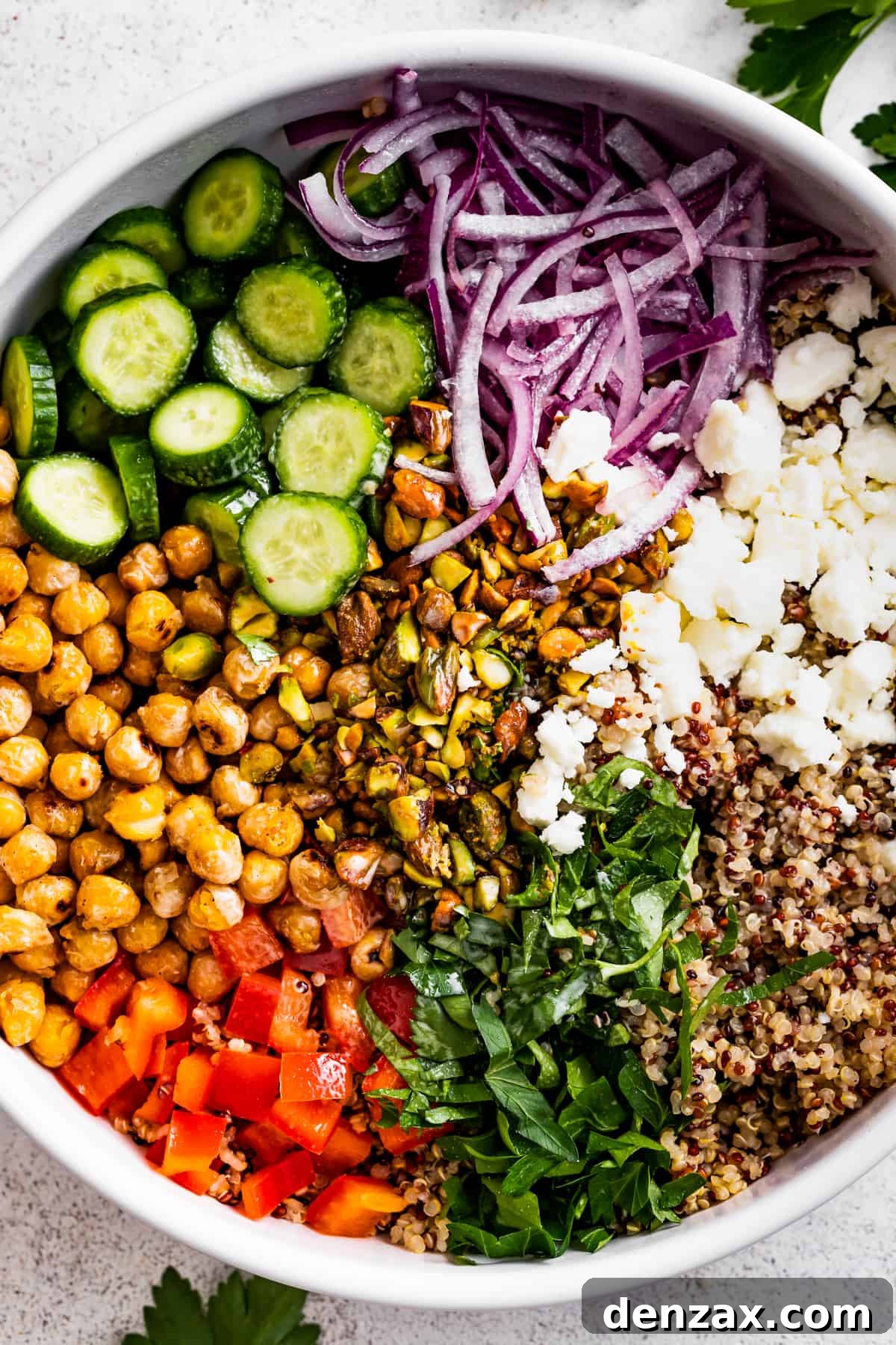 Drizzled with homemade dressing, this Jennifer Aniston quinoa salad with crunchy chickpeas is super light and refreshing. A large bowl with sliced cucumbers, sliced red onions, chickpeas, diced red bell peppers, herbs, tri-colored quinoa, and crumbles of feta cheese.