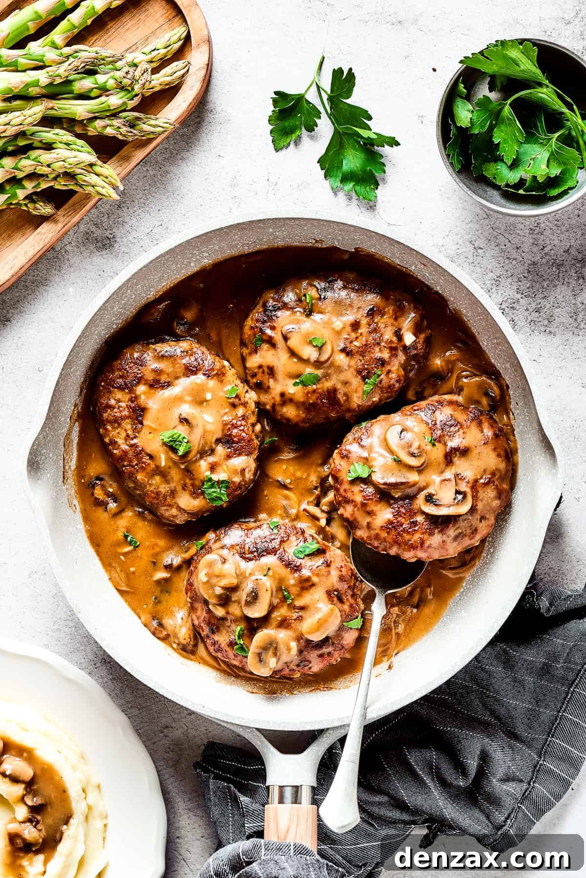 Four salisbury steaks in gravy, in a skillet. Vegetables, herbs, and a cloth napkin are arranged around the skillet.