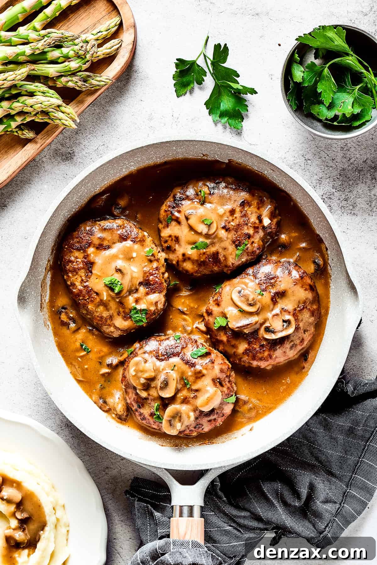 Four salisbury steaks in gravy, in a skillet. Vegetables, herbs, and a cloth napkin are arranged around the skillet.