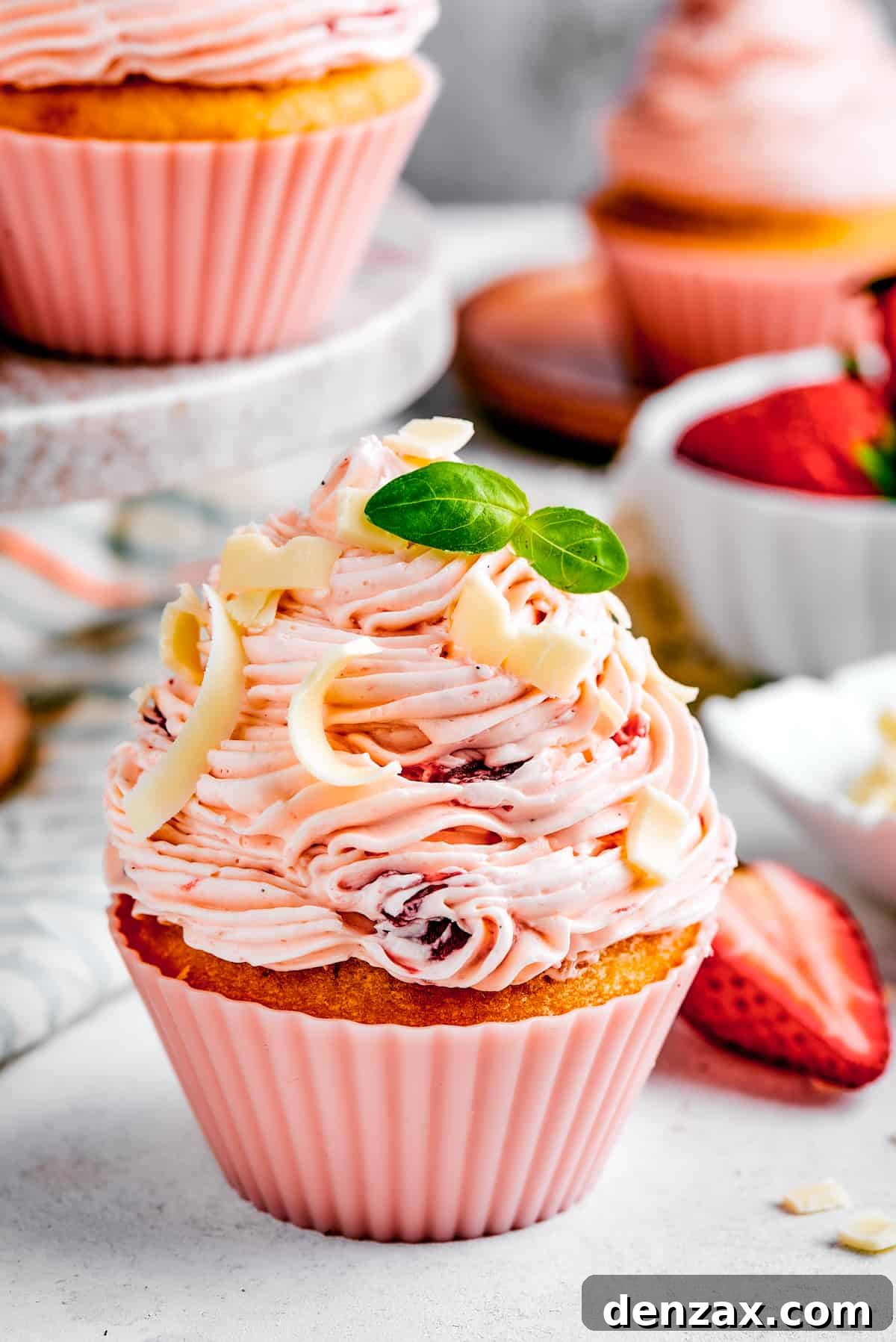 A strawberry cupcake in a pink cupcake liner with a tiny sprig of mint on top. More cupcakes are visible in the background of the shot.