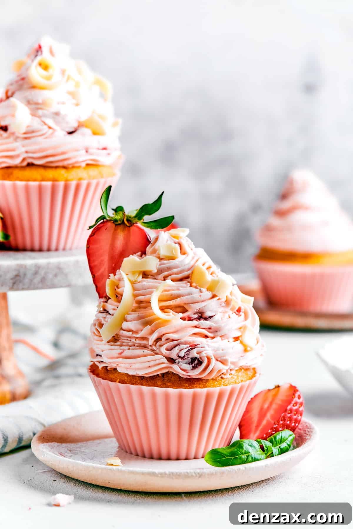 A strawberry cupcake in a pink cupcake liner with a tiny sprig of mint on top. More cupcakes are visible in the background of the shot.