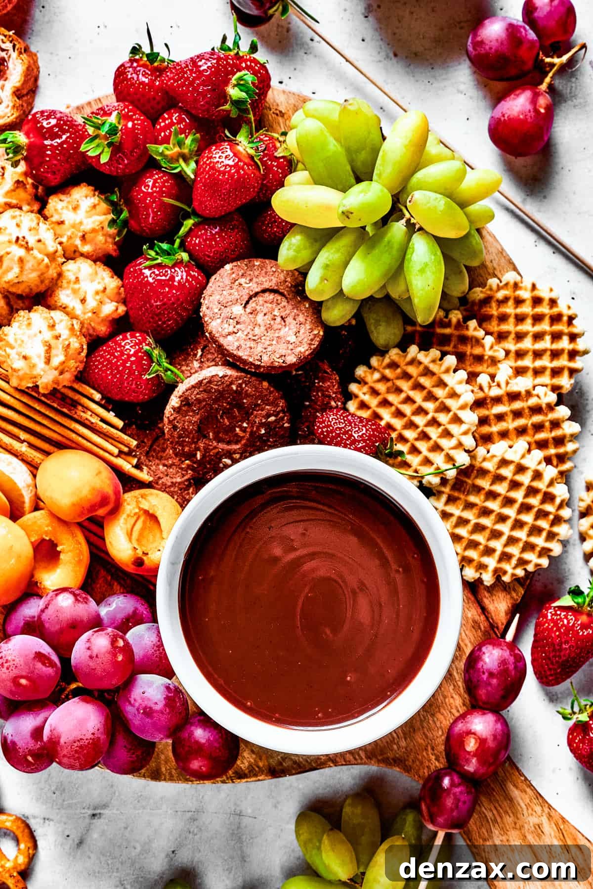 A dessert board with fruit, waffles, pretzels, and a bowl of fondue in the center.