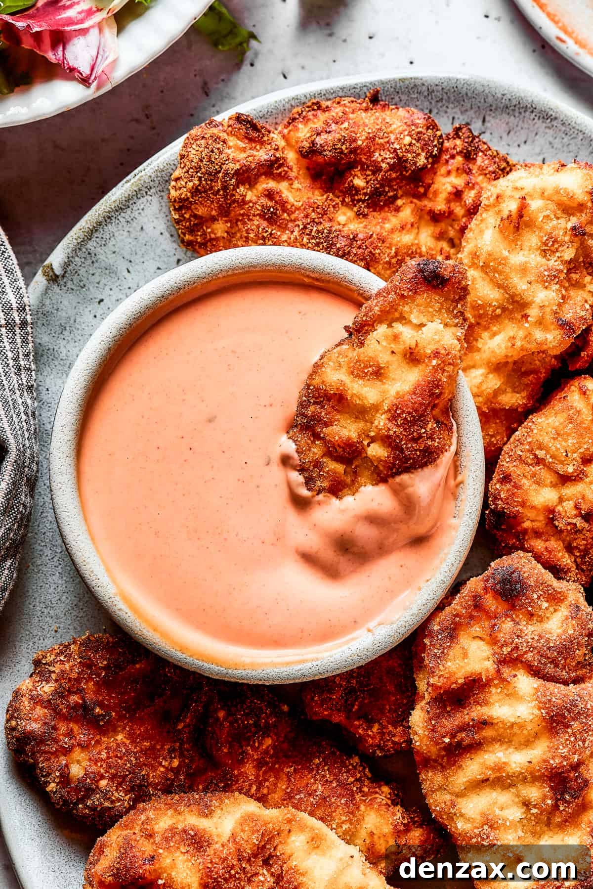 Yum Yum Sauce is a scrumptious, creamy condiment that adds a burst of flavor to any dish. You'll love this sweet, tangy Japanese sauce! A platter of chicken strips with one piece dipped in a bowl of sauce.