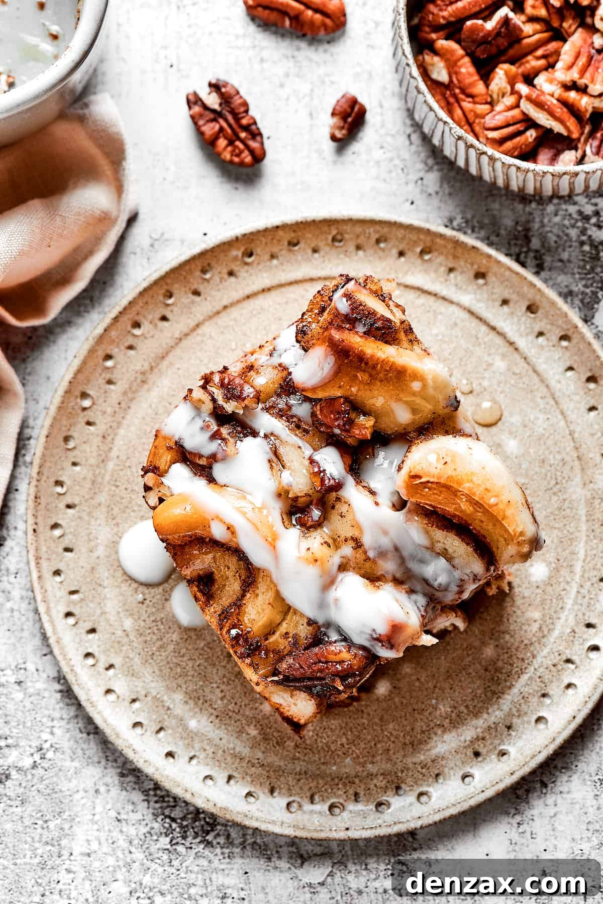 These easy and celebratory cinnamon roll casseroles are rich, creamy, and buttery, and packed with sticky and sweet maple syrup and pecans. Overhead view of a slice of cinnamon roll casserole on a plate, next to a bowl of pecans