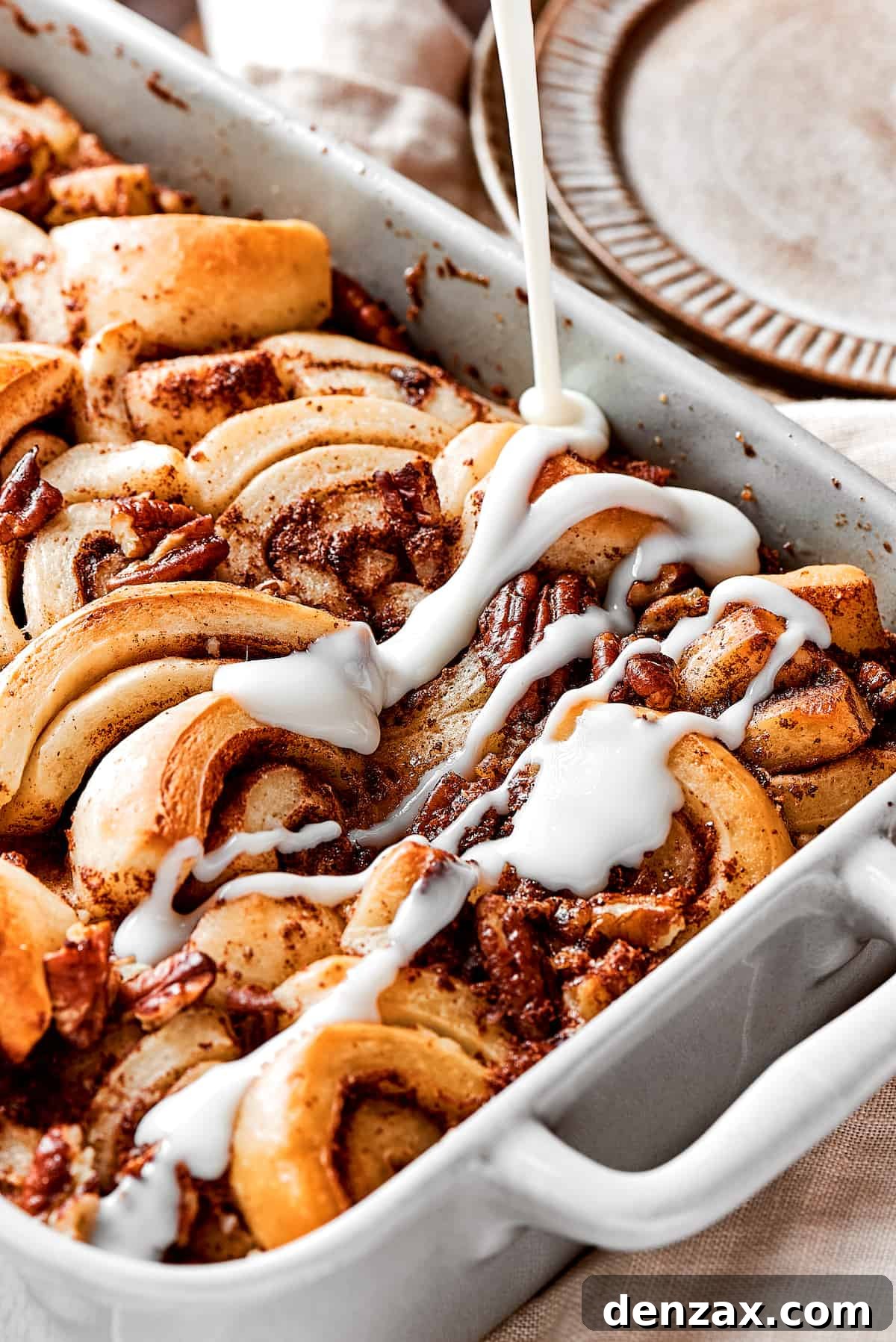These easy and celebratory cinnamon roll casseroles are rich, creamy, and buttery, and packed with sticky and sweet maple syrup and pecans. A cinnamon roll casserole with icing being drizzled over the top