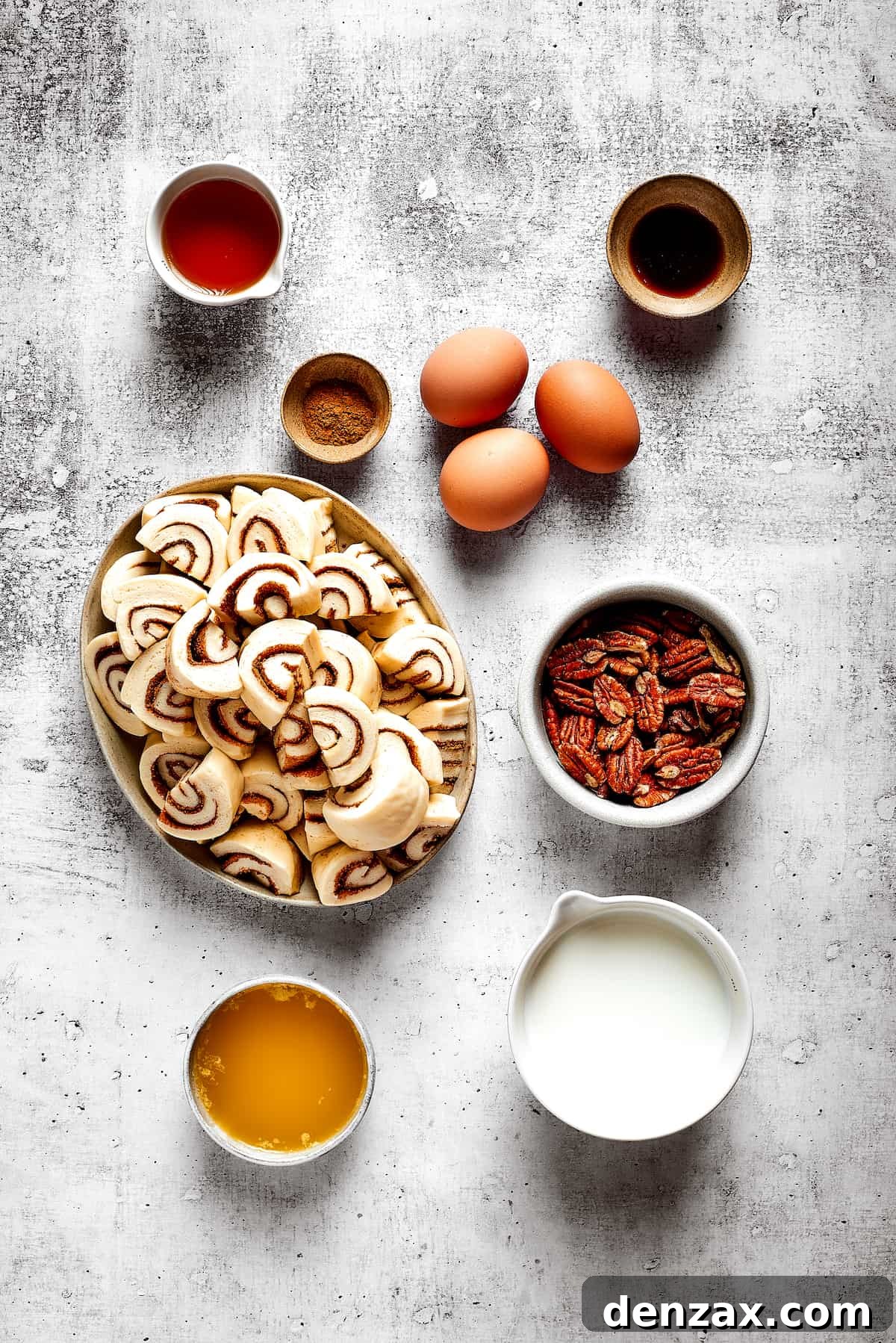 These easy and celebratory cinnamon roll casseroles are rich, creamy, and buttery, and packed with sticky and sweet maple syrup and pecans. Overhead view of the ingredients needed for cinnamon roll casserole: a plate of cinnamon roll pieces, a bowl of milk, a bowl of maple syrup, a bowl of pecans, three eggs, a bowl of cinnamon, a bowl of vanilla, and a bowl of melted butter