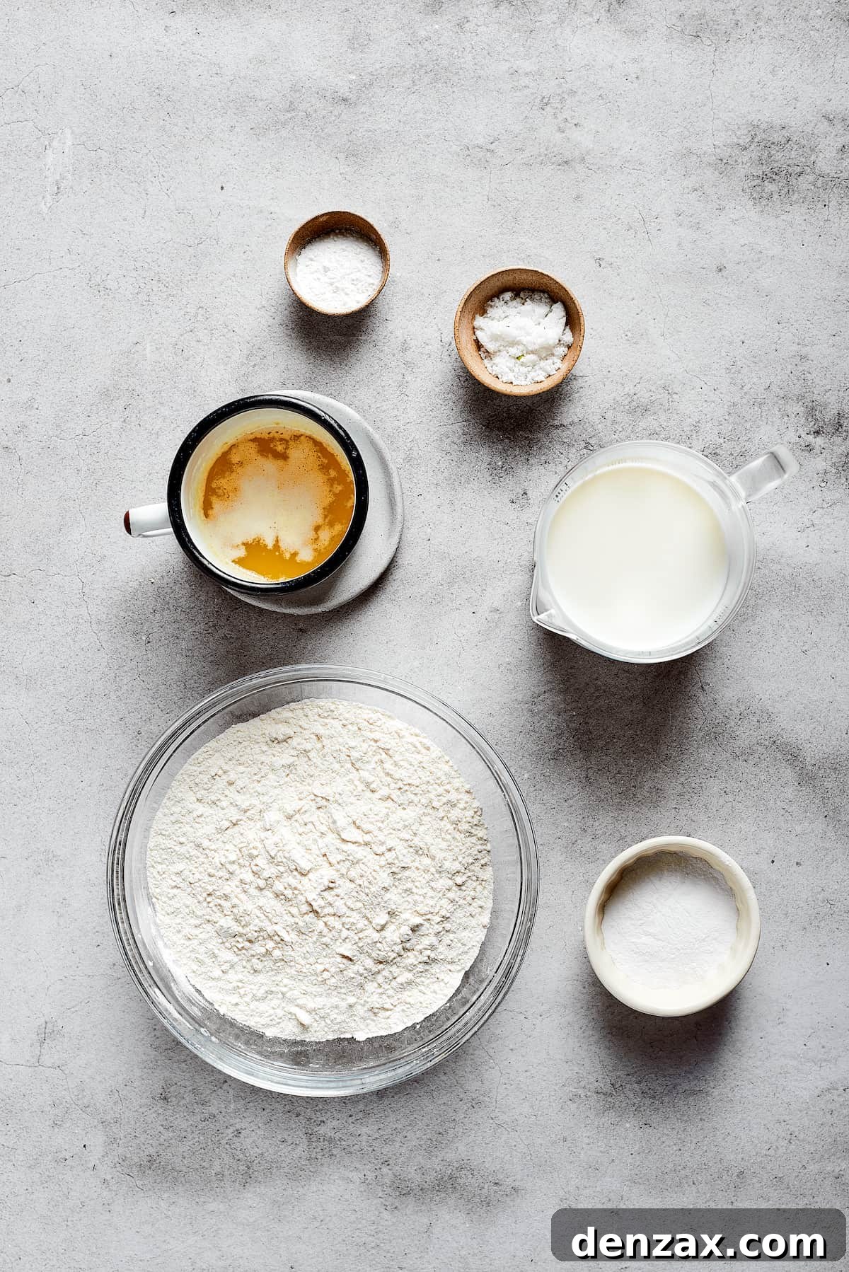 Overhead view of the ingredients needed for butter swim biscuits: a bowl of flour, a cup of melted butter, a pyrex of buttermilk, a bowl of sugar, a bowl of salt, and a bowl of baking powder