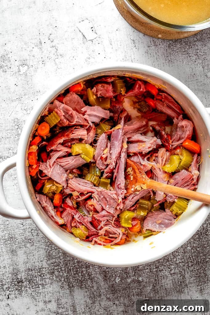 Overhead view of a dutch oven filled with veggies, ham, herbs, and a wooden spoon, next to a pyrex of broth.