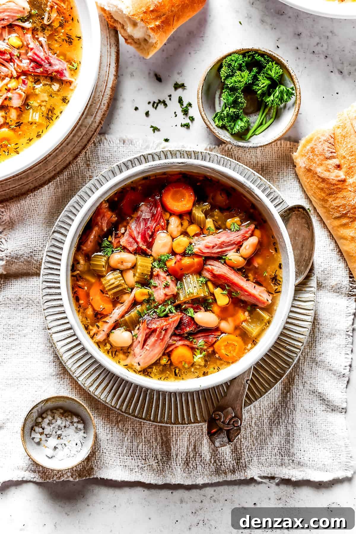 Overhead view of a bowl of ham bone soup with a spoon in it, next to a few pieces of baguette, another bowl of soup, and a bowl of salt.