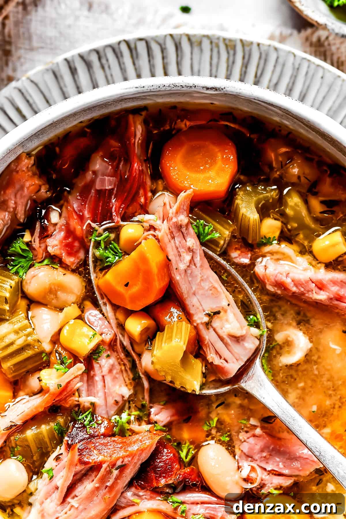 Close up of a spoonful of ham, bean, and veggie soup, above a bowl of the soup.