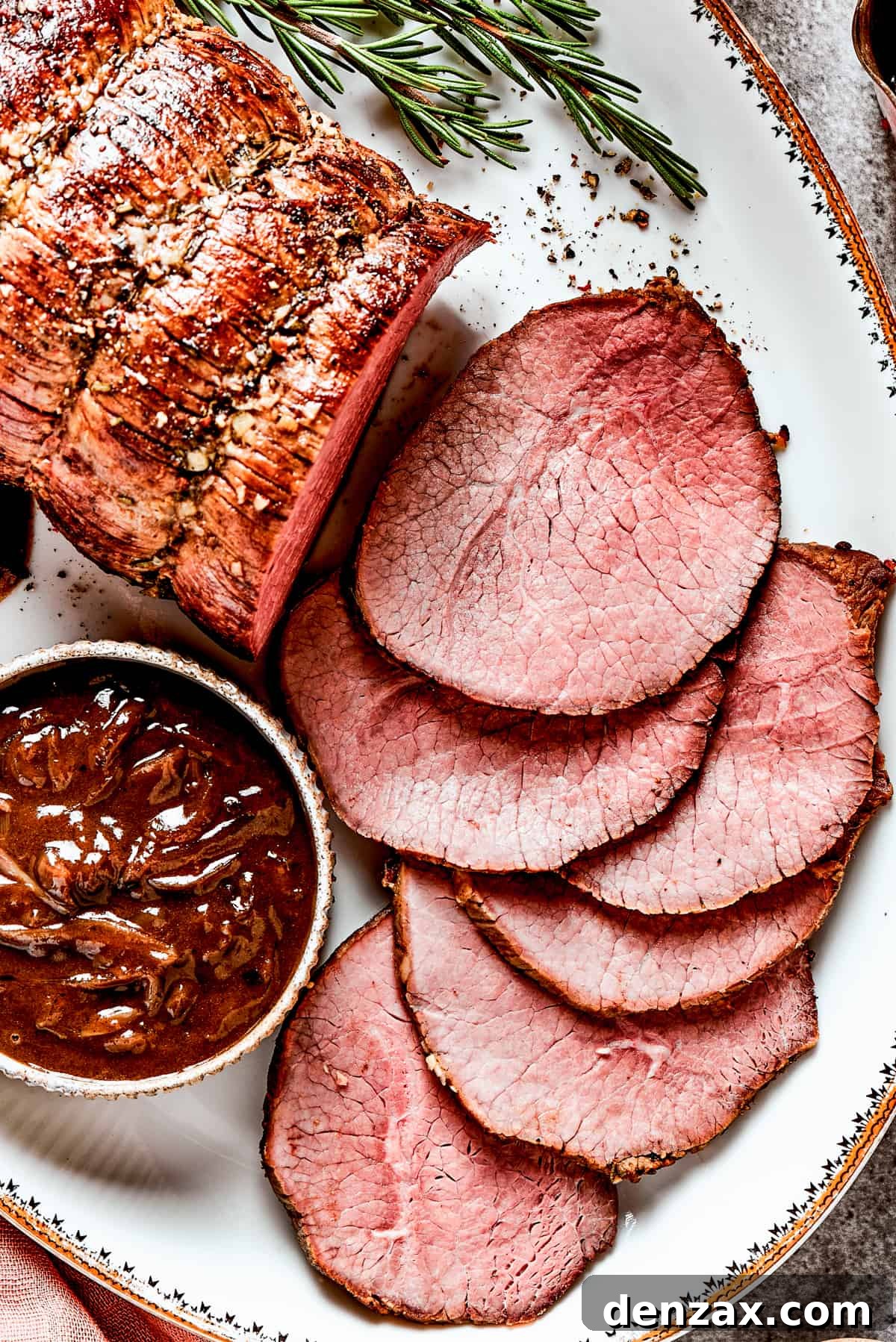 Image of a sliced beef roast served on a white platter with a bowl of gravy set next to it.